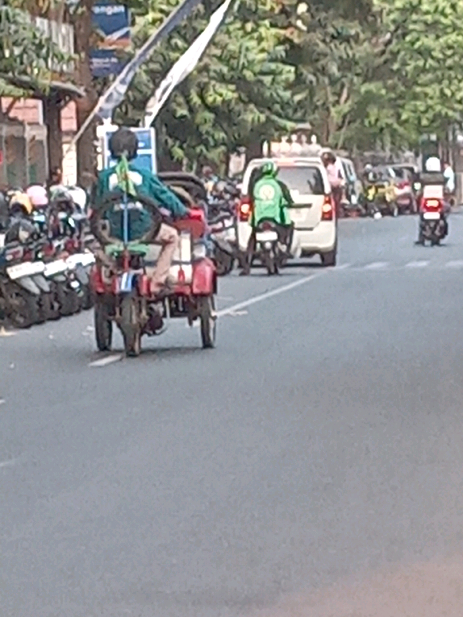 The image depicts a street scene in Yogyakarta, Indonesia, with several vehicles and people. In the foreground, a three-wheeled vehicle, possibly a modified motorcycle or a small cargo vehicle, is seen from the rear. It is a red and white contraption with a driver wearing a green jacket and a dark helmet. The driver appears to be steering the vehicle, which has two wheels at the rear and one at the front, although the front wheel is not clearly visible. The vehicle is loaded with items, including what appears to be a spare tire or wheel on the back.
To the left of this vehicle, a row of parked motorcycles is visible. In the middle ground, a white car is driving away from the camera, with a person in a green jacket, presumably a rider on a motorcycle, partially visible behind it. Further in the background, other vehicles, including motorcycles and a larger van-like vehicle, are present in traffic.
The environment suggests a busy urban street. Trees with lush green foliage line the right side of the road, indicating a relatively warm climate. Buildings are partially visible on the left side of the image, along with street poles and banners, one of which has Indonesian text. The road surface is asphalt, and there are white lane markings visible. The overall lighting suggests it is daytime, with sunlight creating shadows. The image quality is somewhat blurry, making precise identification of all details challenging.