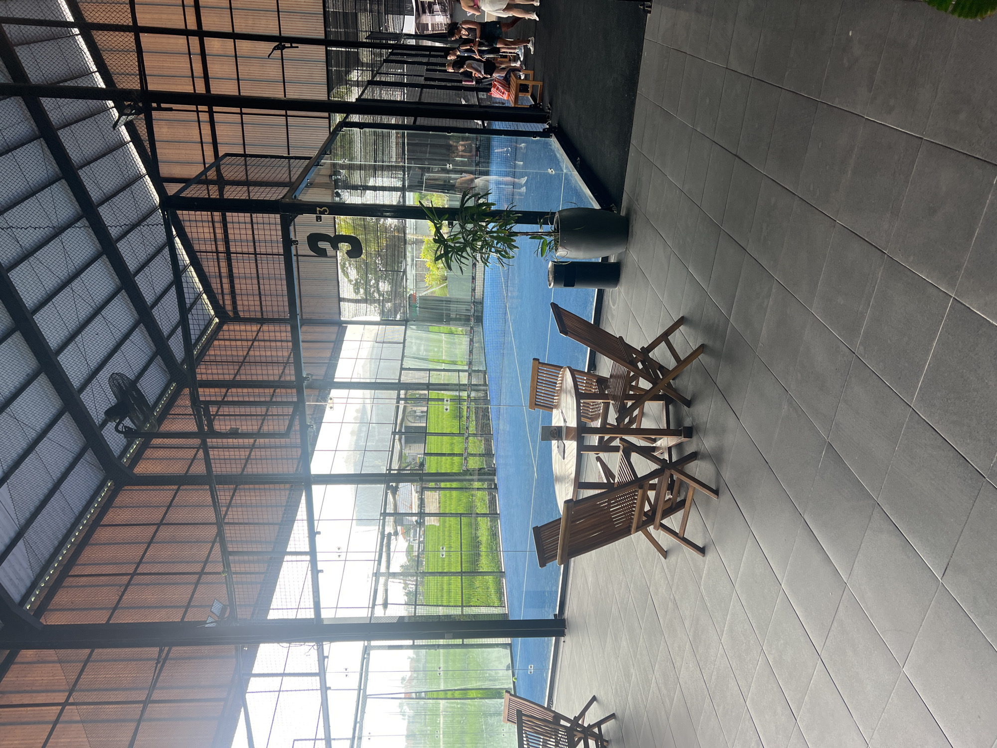 This is a wide, eye-level shot of a brightly lit indoor padel tennis court complex in North Kuta, Indonesia. The court has a blue surface and is enclosed by glass walls. Outside the court, lush green rice fields are visible through the glass. In the foreground, a round wooden table is surrounded by four wooden chairs, set on a dark gray tiled floor. To the right, the court is populated by several people, some standing and others seated on a wooden bench. A potted plant is placed near the glass enclosure. A large black number "3" is visible on the glass wall of the court. The ceiling is constructed with corrugated metal and translucent panels, with exposed metal beams and a hanging fan. The overall impression is one of a modern sports facility with a connection to the surrounding natural landscape.