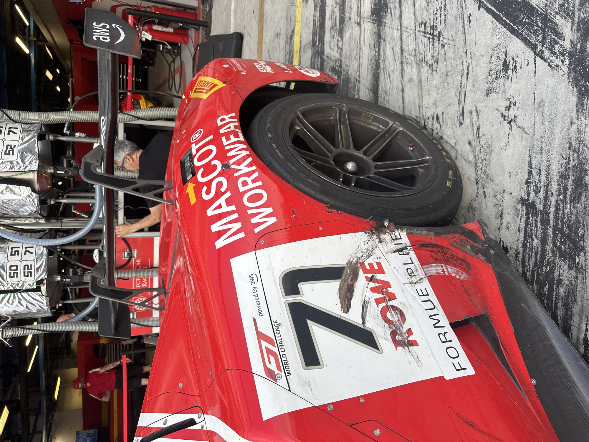 Here's a detailed description of the image:
The image shows the damaged red race car in a garage setting, specifically the pit lane area, based on the presence of the car and crew.
**Subject/Object:**
* The race car is red and white with sponsor logos (MASCOT WORKWEAR, ROWE FORMUE PLAIE, Pirelli, and GT World Challenge) and racing number "71." It shows visible damage (scratches and breakage) on the front passenger side.
* There's an AWS (Amazon Web Services) logo on the wing.
**People:**
* An adult male is visible inside or working on the car within the garage.
**Setting/Environment:**
* The location is likely in a pit lane or garage area associated with a racetrack. This is based on the presence of race equipment, tools, and garage fixtures.
* Given the ROWE FORMUE PLAIE sponsorship it is likely a European track or race event. Given that a tire is resting on what seems to be the floor of the garage it could be in the city of Monza, Italy.
**Actions/Interactions:**
* The race car seems to be undergoing maintenance or inspection after potential damage.
* The man is focused on some task.