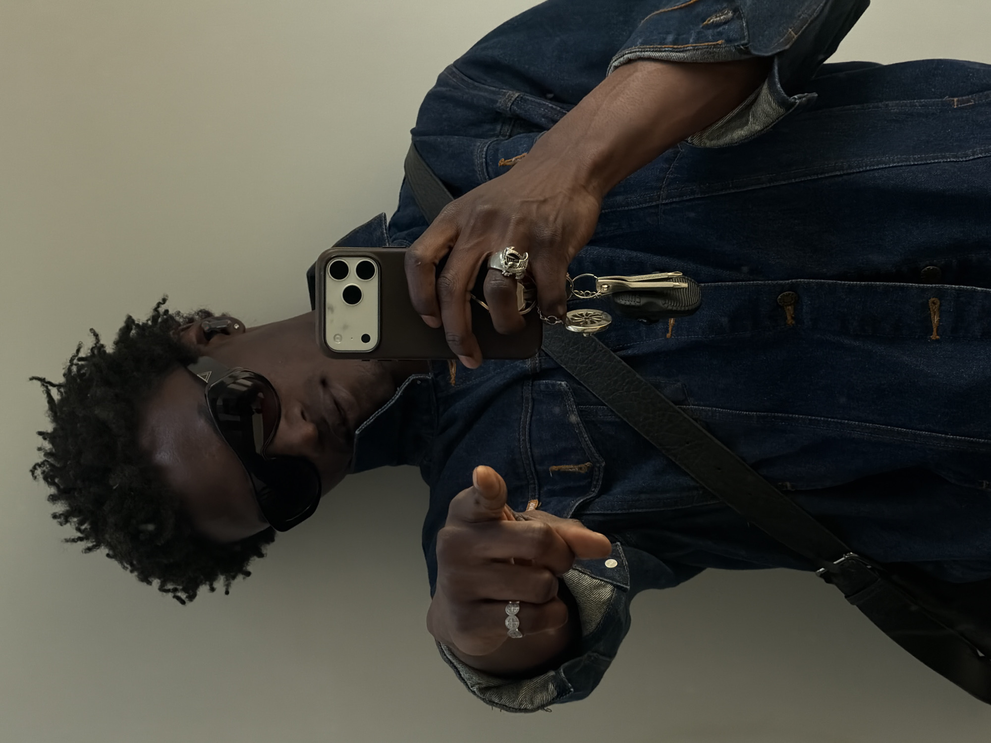 The image captures a mirror selfie of a person with dark skin, wearing a dark blue denim jacket and dark sunglasses. The person is positioned against a plain, light-colored wall. They hold a dark-colored smartphone with a prominent triple-lens camera module in their right hand, with the phone's camera facing towards the viewer. Their left arm is bent, and the index finger of their left hand is pointing directly at the camera, engaging the viewer.
Visible accessories include several rings on the person's right hand: a multi-band ring with decorative elements on the ring finger and a larger, ornate silver ring, possibly skull-shaped or highly detailed, on the middle finger. A set of keys attached to a keychain, which features a circular, compass-like or decorative disc and other components, hangs from the right hand near the phone. A dark strap, likely from a bag, crosses diagonally over the person's chest.
A small, triangular logo is faintly visible on the temple of the sunglasses, though too blurry to identify precisely. Illegible text can also be seen on a black component of the keychain. The overall mood is confident and direct, with the sunglasses obscuring facial expression. The setting appears to be indoors, with no specific weather or time of day cues. The provided context indicates the location is London, United Kingdom.