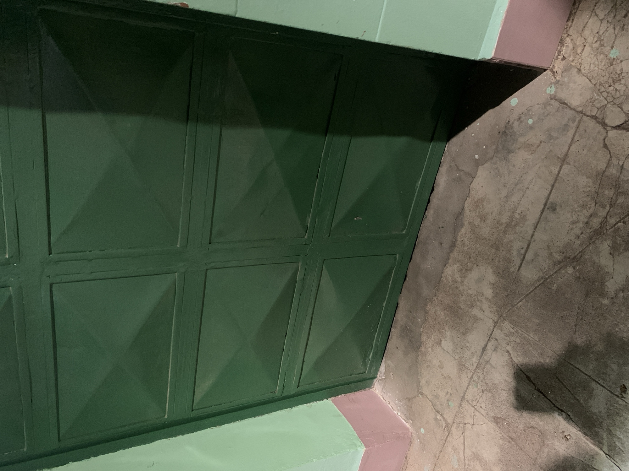 A low-angle shot shows a section of a green metal door with raised square panels. Above the door is a light green wall with a strip of pink paint along the bottom edge. The door is positioned to the left. To the right, the concrete floor has an off-white base color with prominent dark lines creating rectangular shapes. There is also some light green splatter. Cracks are visible across the concrete surface. The scene has moderate lighting. Without additional information, the exact location in Santa Ana, Venezuela cannot be determined.