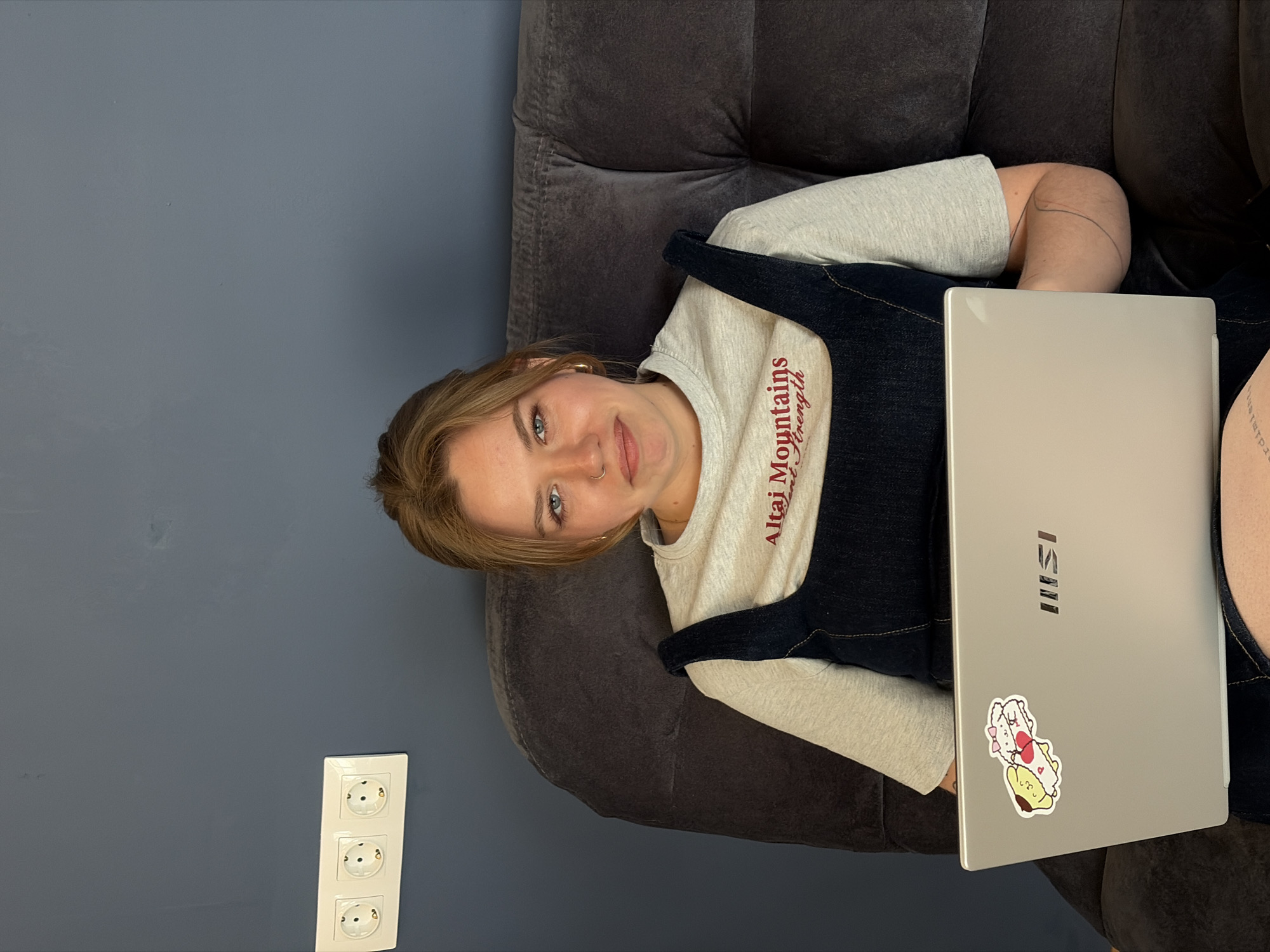 A woman is sitting on a grey couch, looking at the camera while holding a silver MSI laptop on her lap. She is wearing a light grey t-shirt with maroon text on it and a dark blue denim pinafore dress. The wall behind the couch is a dark blue-grey color. On the wall is a white electrical outlet with three sockets. The location is inferred to be in Бараўлянскі сельскі Савет, Belarus.