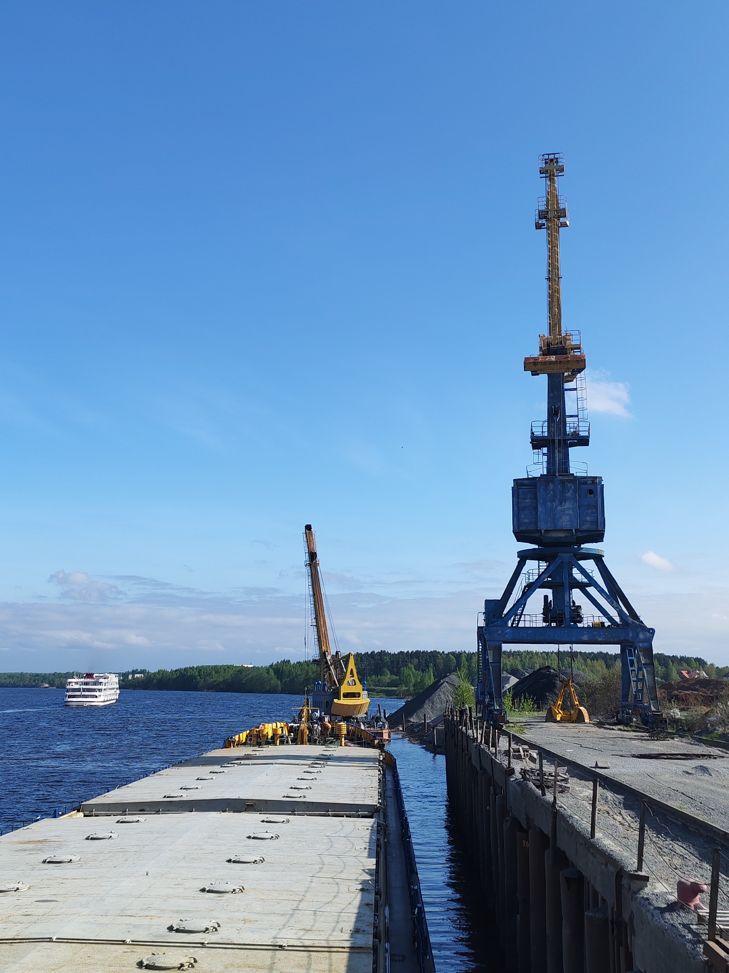 The image captures a scene at the port of Tver, Russia. In the foreground is the deck of a large barge, adjacent to a concrete dock. A narrow waterway separates the barge from the dock.
Two large cranes dominate the right side of the image. The crane closer to the viewer is dark blue and constructed of heavy steel. It stands on a concrete base, extending upwards into the clear blue sky. Behind the blue crane, a yellow crane is visible, also working on the dock.
Piles of gravel are located along the dock, suggesting the cranes are used for loading and unloading materials.
In the background, a river or wide waterway is visible, with a white passenger ship sailing in the distance. The opposite bank of the waterway is covered with green trees, indicating a natural environment bordering the industrial area. The sky is blue with very few clouds. No people are clearly visible in the scene.