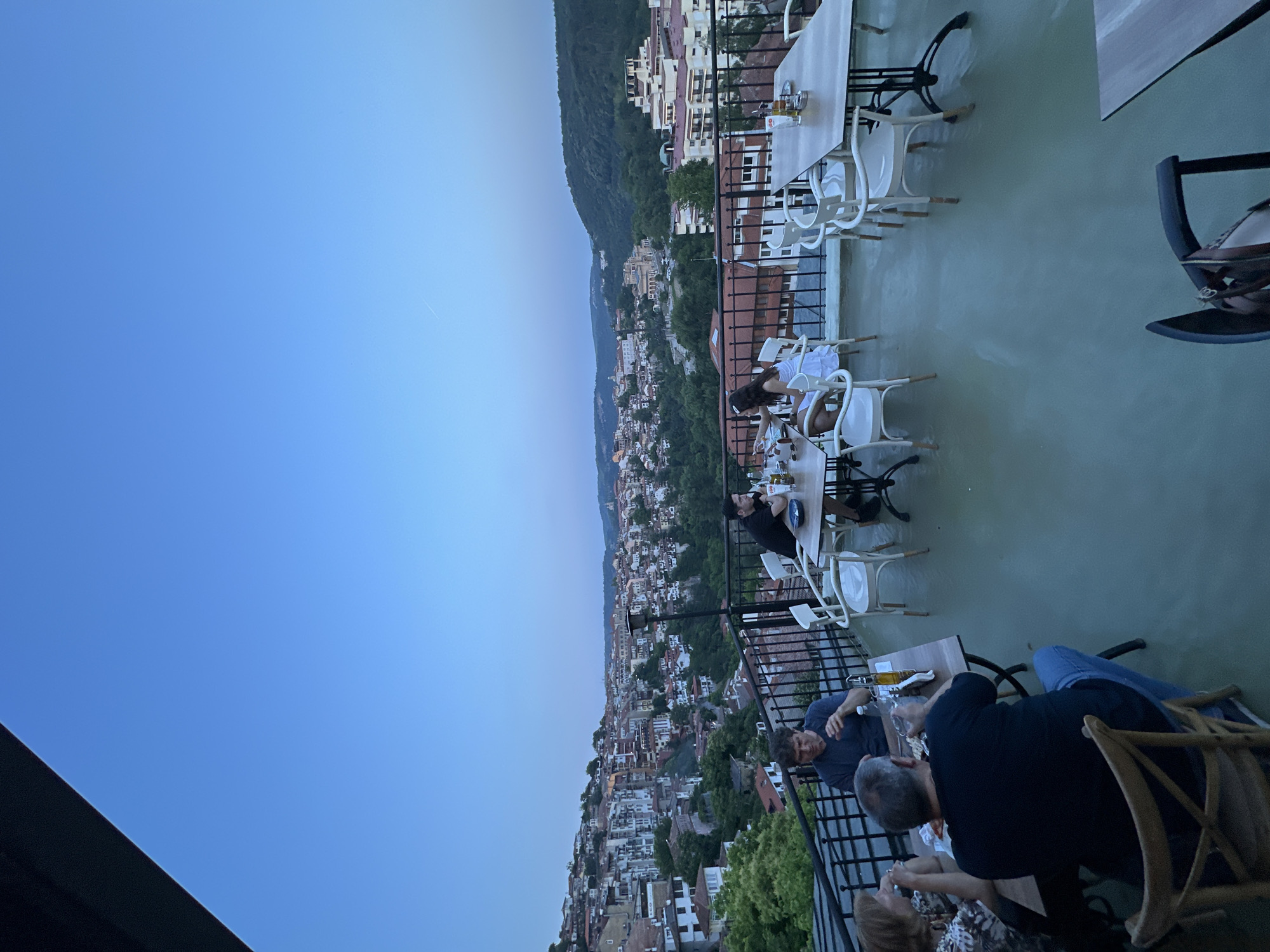 An outdoor restaurant terrace provides a panoramic view of Veliko Tarnovo, Bulgaria, at dusk. The sky is a clear, deep blue with a faint aircraft contrail visible in the upper right.
In the foreground, the terrace floor is a light greenish-grey. A dark metal railing with horizontal bars encloses the dining area, offering an unobstructed view of the city. Several tables, with light wooden tops and dark metal bases, are arranged across the terrace. Seating includes white chairs, some with armrests, and natural light brown wooden cross-back chairs.
On the left, a group of four people is seated around a table. An older woman with light brown hair, wearing a patterned top, is looking towards a man with grey hair, dressed in a black t-shirt and blue jeans, who is holding a glass with his back partially to the camera. Opposite him, a man with dark, curly hair in a dark blue t-shirt gestures with his hand. Their table holds bottles (one olive oil), glasses, and a white bowl of snacks.
In the mid-center, a man with dark hair and a beard, wearing a black t-shirt, faces a woman with long dark hair in a white dress, seated at a separate table. Their table has bottles, glasses, and plates. Further right, a man with dark hair, wearing a black t-shirt, sits alone at a table, leaning his chin on his hand and looking out at the view. An unoccupied table with white chairs is to his right. A light brown handbag rests on a chair in the lower right corner. A tall, dark pole, possibly a heater or light fixture, stands near the railing on the terrace.
The midground showcases the densely built cityscape of Veliko Tarnovo, characterized by buildings with red-tiled roofs and light-colored facades cascading down steep, forested hillsides. Trees and green vegetation are interspersed throughout the urban fabric. Prominent buildings, including what appears to be a larger, lighter-colored public building or church, are visible among the residential structures.
In the background, verdant hills extend into the distance under the twilight sky. The overall lighting is dim and ambient, indicative of the evening.