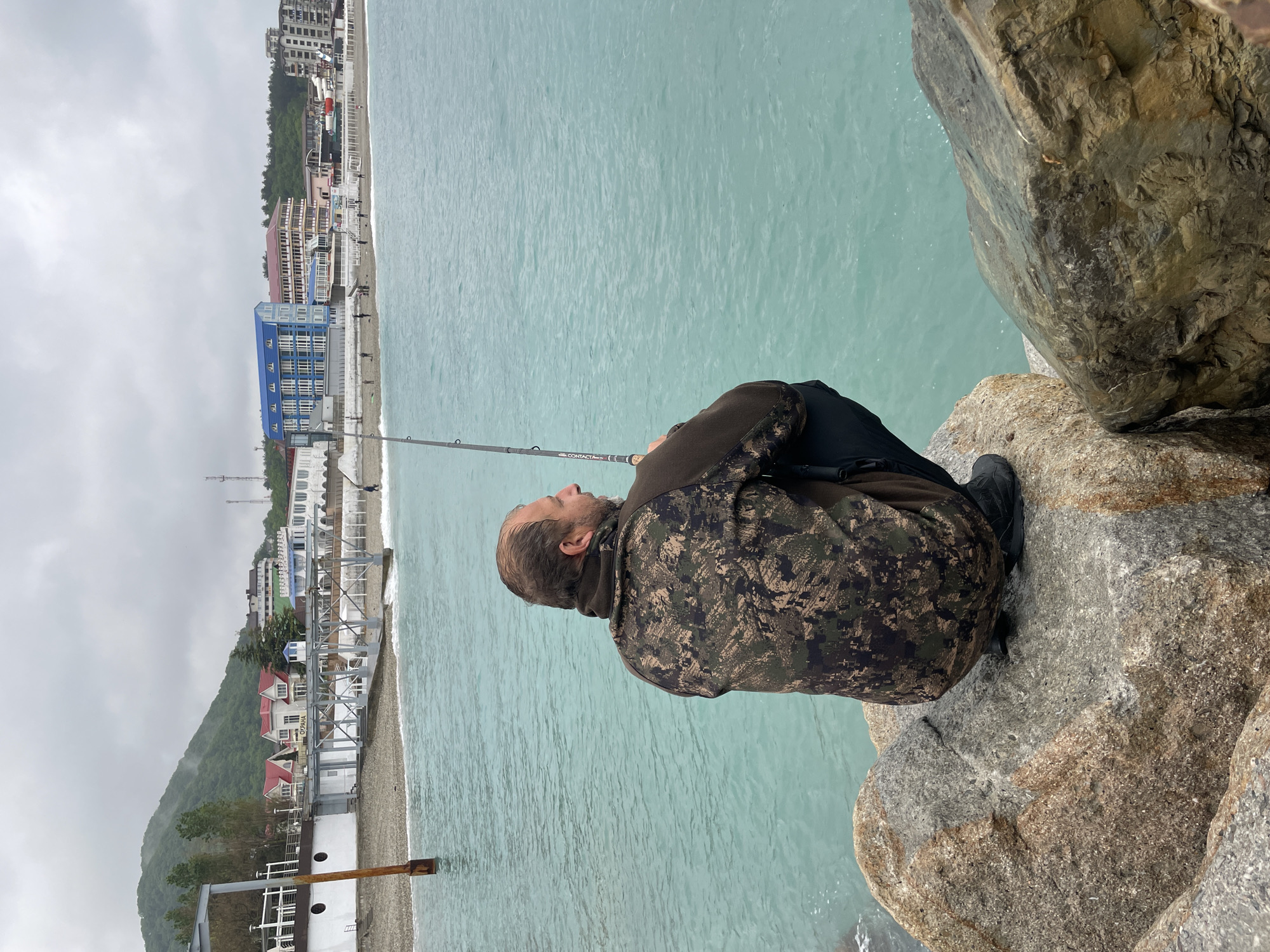 A man wearing a camouflage jacket, dark pants, and black shoes sits on a rocky outcropping, holding a fishing rod over turquoise water. The setting is Olginka, Russia. A shoreline and buildings are visible in the distance under an overcast sky.