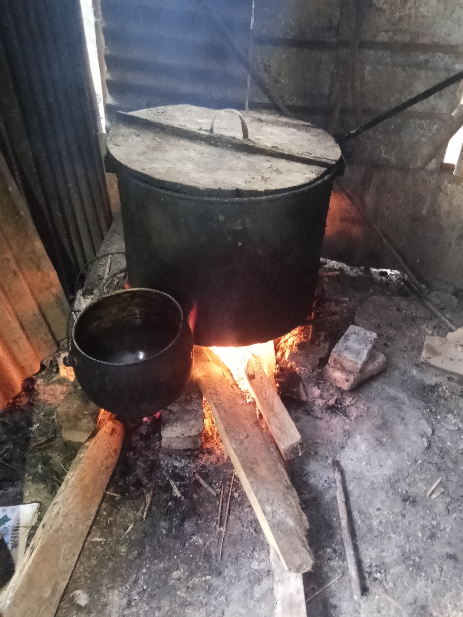 The image depicts an outdoor cooking scene. Two large, dark pots are present, one significantly larger than the other. The larger pot is covered with a lid and is situated over a fire, with visible flames and smoke emanating from underneath. It appears to be made of metal. To its left, a smaller, cauldron-like pot is open and also appears to be made of metal. The ground is covered with ash, scattered pieces of wood, and some bricks, suggesting a makeshift cooking area. The surroundings appear to be rustic, with corrugated metal sheeting visible in the background, possibly forming a wall or shelter. The overall impression is of a traditional or rural cooking setup. The image does not contain any specific landmarks or contextual clues that would indicate the city of Makale, Indonesia.
