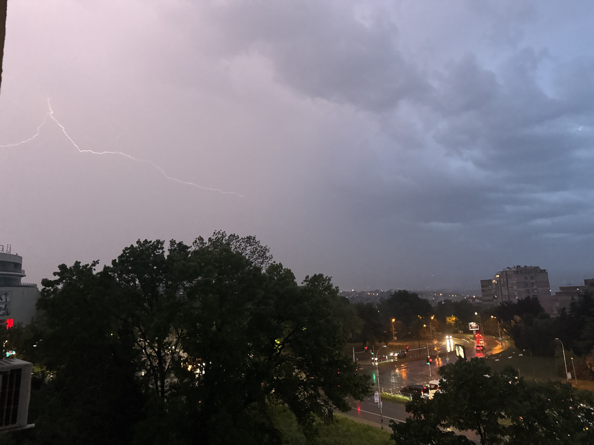 A wide-angle shot depicts a stormy cityscape, possibly Varna, Bulgaria, during a lightning storm. The sky is dominated by dark, ominous clouds, with a bolt of lightning extending across the left side of the frame. Below the sky, the scene transitions to a view of urban life. Buildings are scattered throughout, interspersed with dense, green trees. Streetlights illuminate a road below, reflecting off the wet pavement. Red and white car lights suggest ongoing traffic. The photograph is taken from an elevated viewpoint, possibly a high-rise building, offering a broad perspective of the city in stormy weather.