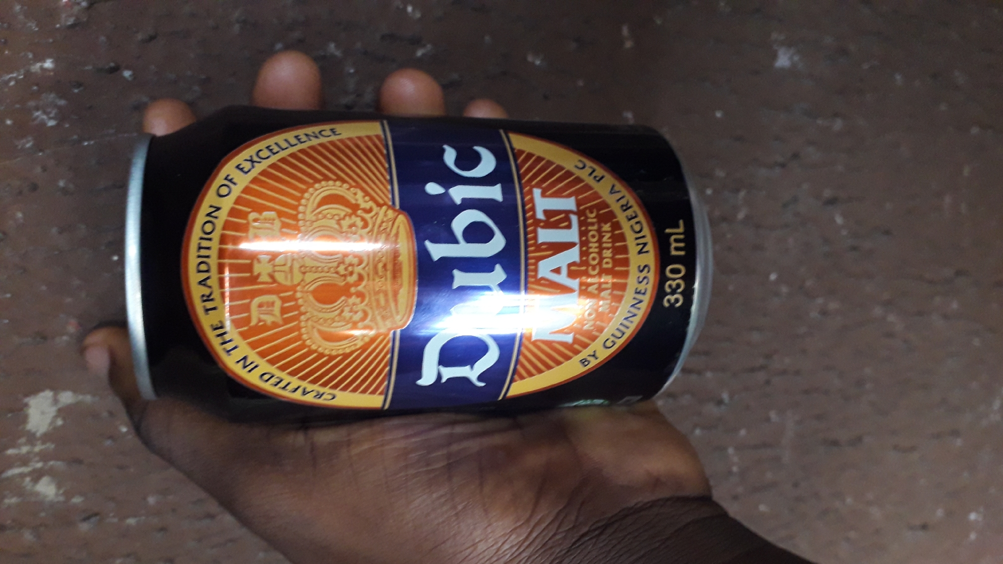 A dark-skinned hand holds a can of "Dubic Malt Non-Alcoholic Malt Drink." The can is mostly black, with gold and blue accents. It is marked "Crafted in the Tradition of Excellence" and "By Guinness Nigeria PLC, 330 mL." The background is an indistinct, neutral-colored surface. The location is Nkpor, Nigeria.