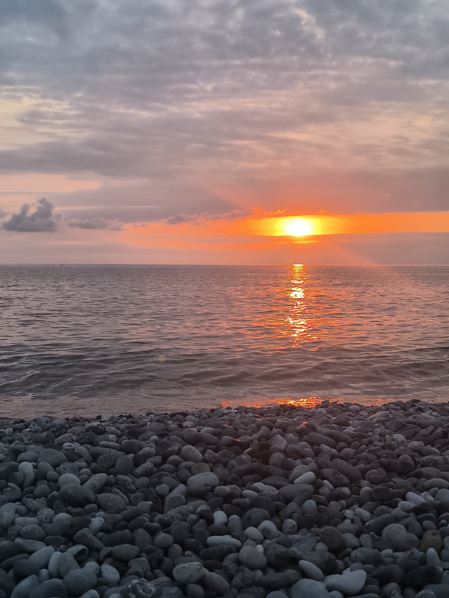 The image captures a tranquil sunset over the Black Sea, as viewed from a pebble beach in Batumi, Georgia. The sky is a canvas of soft grays and pinks, with a bright orange sun low on the horizon, casting a shimmering reflection on the gently rippling water. The foreground is densely covered with smooth, gray and white pebbles of varying sizes, creating a natural, textured border to the scene. The water is calm, with subtle waves lapping at the shore. The overall impression is one of peace and natural beauty. There are no people or man-made objects visible in the frame, focusing entirely on the serene coastal landscape.