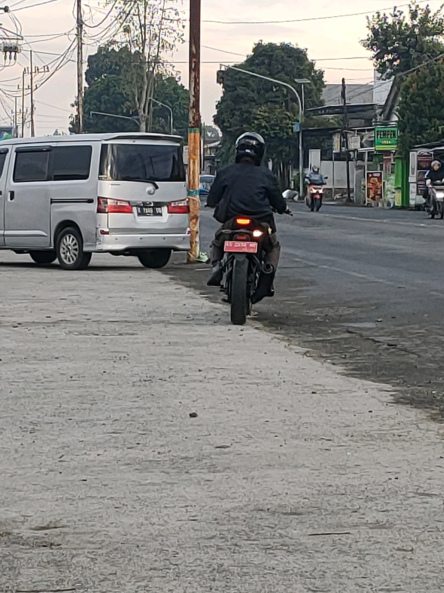 The image captures a street scene in Kanigoro, Indonesia, under an overcast sky. In the foreground, a person is riding a dark-colored motorcycle with red taillights. The motorcycle has a red license plate that is difficult to read. The rider is wearing a black jacket and a black helmet. To the left of the motorcycle, a silver minivan is parked on the side of the road. The minivan has tinted windows and a rear spoiler. The road surface appears to be asphalt, with some debris and discoloration. In the background, there are trees, utility poles with numerous wires, buildings, and other vehicles, including another motorcycle and a car further down the road. A green sign with "PEMPEK" is visible on a building. The overall impression is a typical day on a street in a populated area.