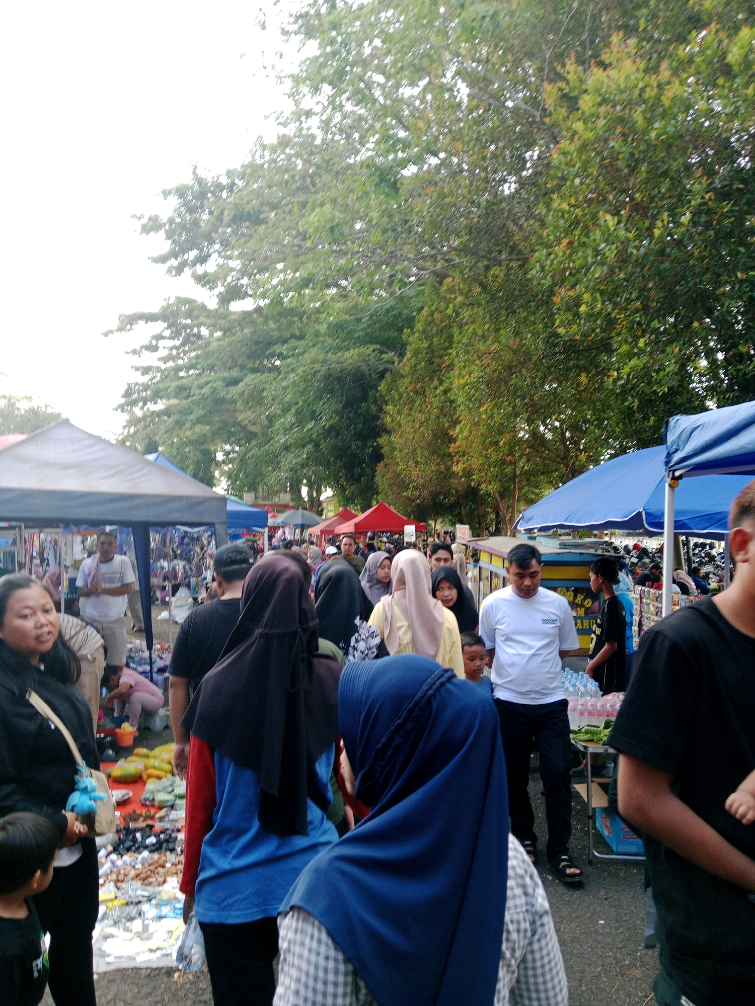 The image depicts a crowded outdoor market scene, likely in Ciamis, Indonesia, given the provided context. A variety of people are walking through narrow aisles between stalls. The stalls are covered by awnings in various colors, including white, blue, and red, and display a range of goods, such as clothing, bags, and possibly food items. The ground is paved or dirt. In the background, lush green trees line the market area, with the sky appearing overcast or bright due to high exposure. The overall atmosphere suggests a bustling marketplace with people engaged in shopping and socializing.