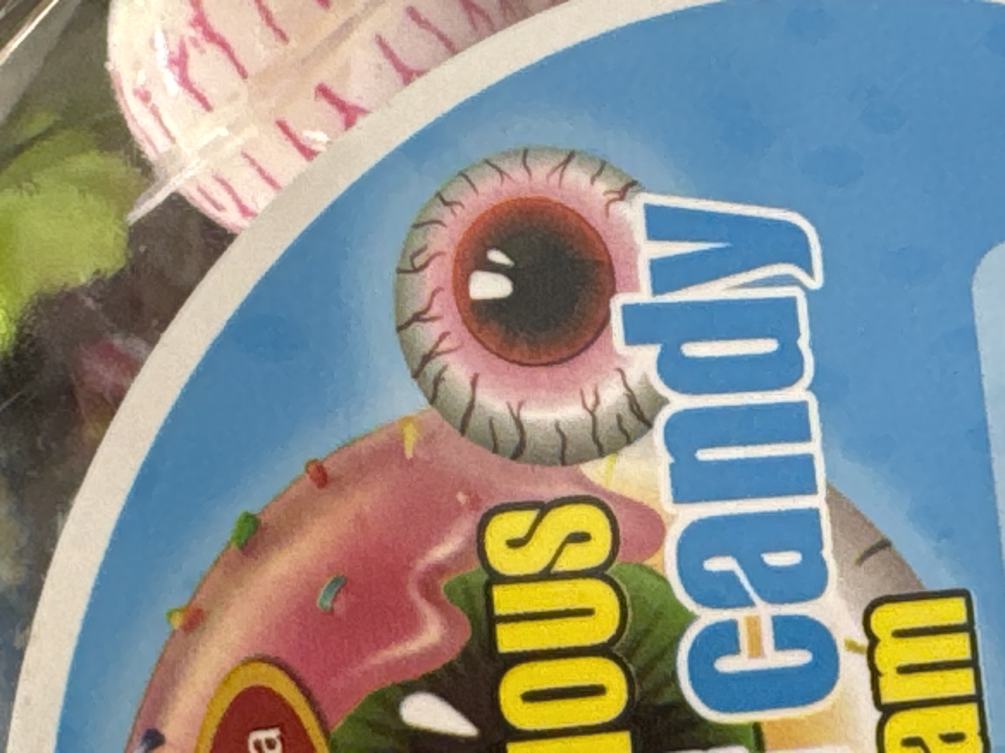 This close-up image captures a portion of a product label, likely for candy, featuring vibrant illustrations and some actual candies in the background. The main focus is on the label's design, which includes an exaggerated illustration of an eyeball with a reddish-brown iris, a dark pupil, and prominent red veins. Adjacent to the eyeball, part of a pink-frosted donut adorned with colorful sprinkles is visible.
In the blurred background, several candies are partially visible, including what appears to be a white and pink striped lollipop or round candy and a bright green gummy-like candy.
The label features prominent text. The word "candy" is clearly visible in large blue letters with a white outline, oriented vertically on the right side of the frame. To the left of "candy," part of another word is visible in yellow letters with a white outline, reading "SNOUS" when interpreted vertically. Given the illustrative style of the eyeball, this yellow text is likely the lower portion of the word "MONSTROUS," suggesting a product name like "MONSTROUS CANDY."
The overall scene suggests a novelty or Halloween-themed sweet treat. No specific activity or event is taking place. The lighting appears bright and even, typical of an indoor setting.