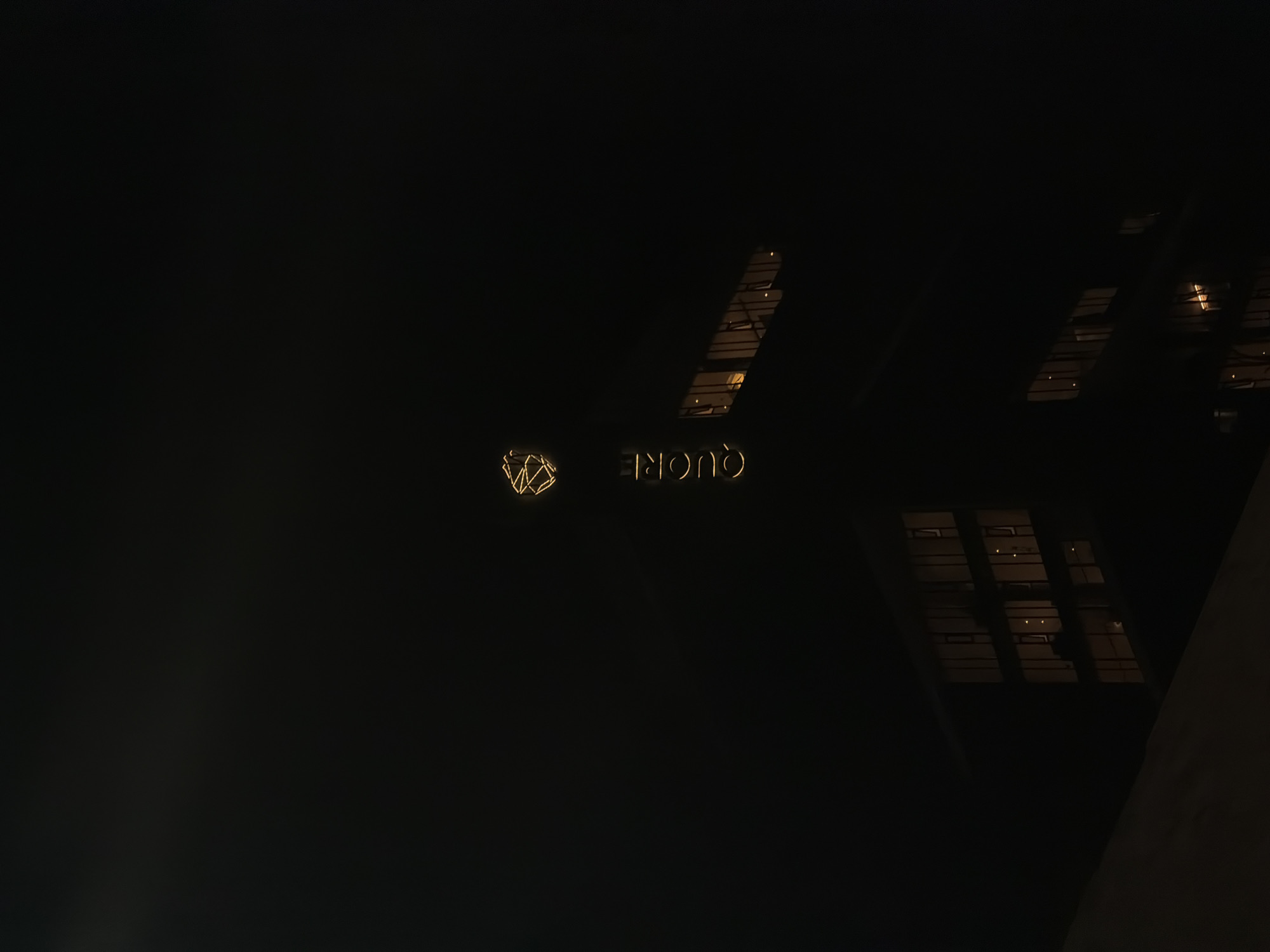 The image captures a predominantly dark scene, suggesting nighttime or a very dimly lit interior. On the left side, a glowing, golden geometric symbol is visible, appearing as a complex, faceted, and abstract shape formed by interconnected lines. To its right, also illuminated in a golden hue, is the inverted word "QUORE". The text and symbol appear to be part of an illuminated sign.
In the upper right quadrant and extending towards the lower right corner, there are reflections or glimpses of warm, yellowish light through a distinct gridded structure, indicative of windows in an adjacent or surrounding building. A dark, diagonal architectural element occupies the bottom right corner of the frame. No individuals are present.
The scene conveys a sense of modernity and subtle illumination against a dark backdrop. Given the location context of Puebla, Mexico, this image likely depicts the nighttime exterior or a specific architectural detail of a building, such as a hotel, restaurant, or commercial establishment, with its distinct branding. The focus is on the glowing signage and the interplay of light and shadow in the dark environment.