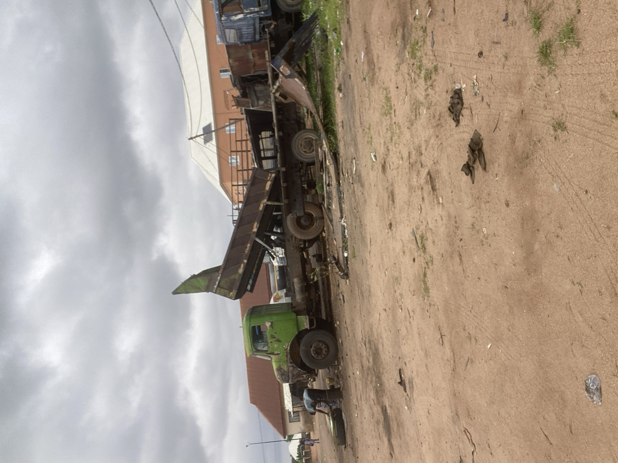 In Gbaza, Nigeria, a scene of disrepair is depicted with two large trucks as the primary subjects. A green dump truck, seemingly inoperable, is positioned on the left side of the frame. A man, wearing a blue shirt and dark pants, is visible in front of the green truck, engaged in an action that appears to be maintenance or repair, possibly working on a tire. A blue truck, also appears to be in a state of disuse and disrepair, is partially visible to the right of the green truck. The trucks are situated in an outdoor, unpaved area, with loose dirt and scattered debris on the ground. Vegetation, including grass, is growing around the trucks and in patches throughout the scene. In the background, buildings with orange walls and white roofs suggest a semi-urban or rural setting. The sky above is overcast with gray clouds. The overall impression is one of abandonment and neglect of heavy machinery in a utilitarian or informal industrial area.