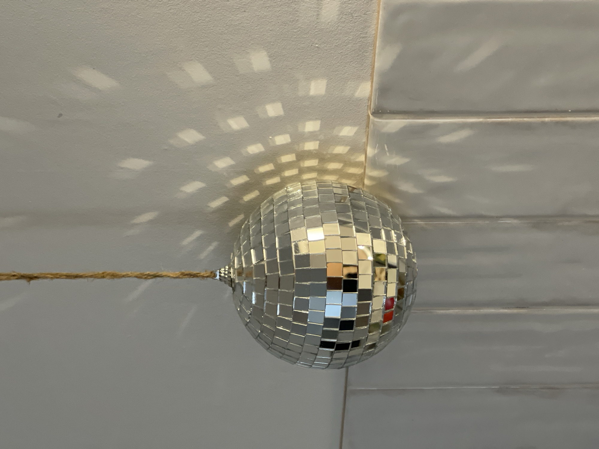 A disco ball hangs from a twine string against a tiled white surface, projecting fragmented light patterns onto the surface. The lighting conditions indicate a light source directed towards the disco ball, causing reflections on the tiles. The tiled surface appears to be a wall or ceiling. The scene lacks the presence of any people. There is no indication in the image to connect it to Izhevsk, Russia.