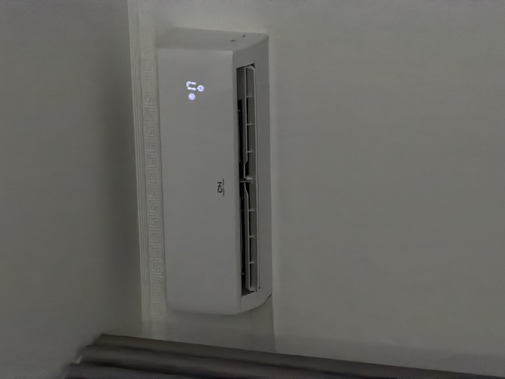 A white air conditioning unit is mounted high on a plain, light-colored wall. The unit has a sleek, modern design with horizontal vents. A small digital display on the right side of the unit shows the temperature as "17" and a circular icon that may represent a fan or power setting. The logo "CH" is visible on the left side of the unit. To the left of the air conditioner, a portion of a grey curtain is visible. The wall features a decorative border or trim just above the air conditioner. The overall scene appears to be indoors, likely in a residential or office setting, in the city of Bila Tserkva, Ukraine.