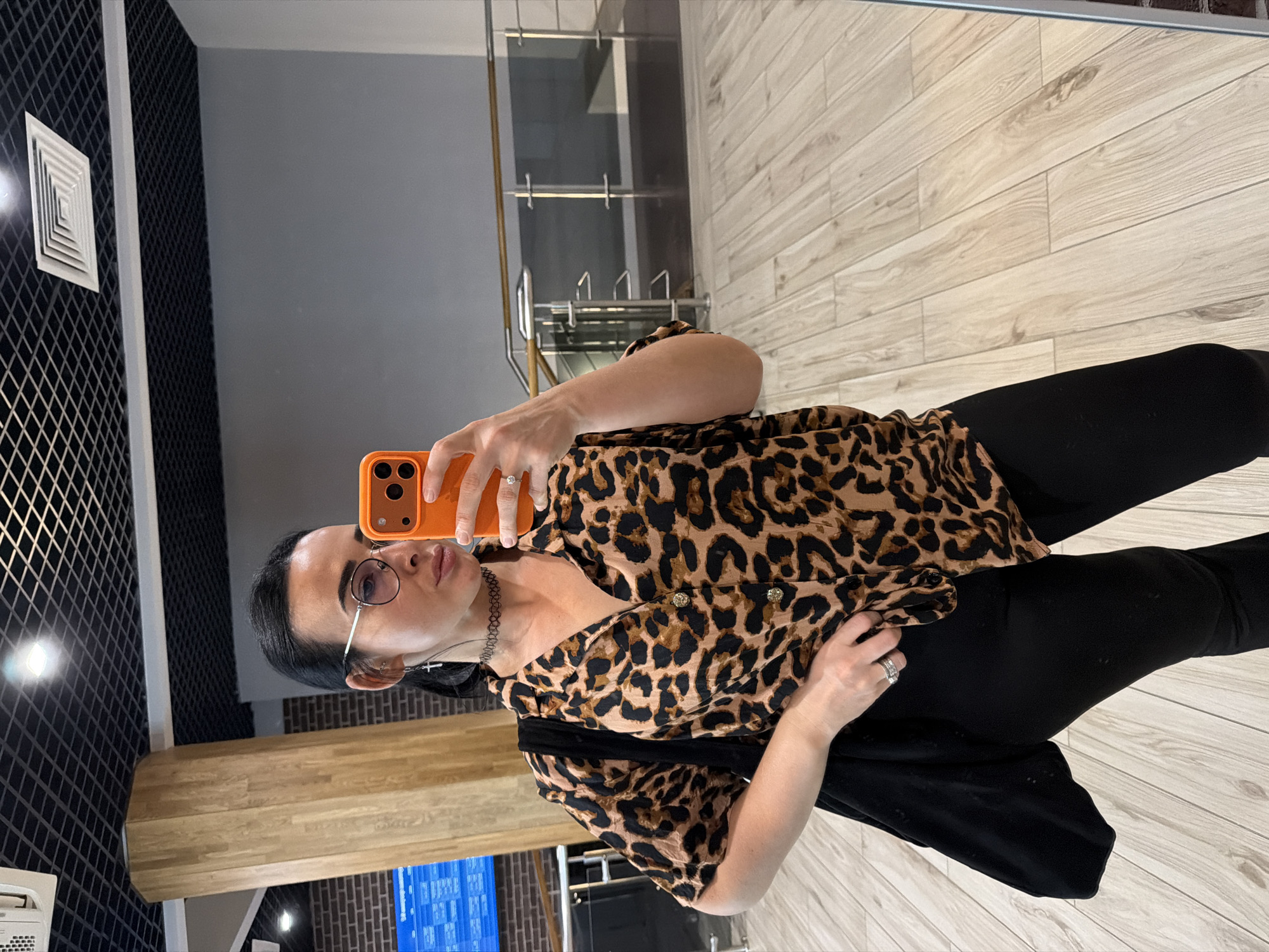 A woman is depicted from the mid-chest down, capturing a mirror selfie indoors. She has dark hair styled back and is wearing round-frame spectacles, a black choker necklace, and a silver cross earring on her left ear. Her attire consists of a short-sleeved, collared shirt featuring a brown and black leopard print pattern, and black trousers. A black fabric shoulder bag is worn over her left shoulder. She holds an orange-cased smartphone in her right hand, angled to photograph her reflection. Visible jewelry includes multiple rings on her fingers, with a prominent ring on her right ring finger. The indoor environment features a light-colored wooden plank floor and a black open-grid suspended ceiling equipped with recessed square lights and air conditioning vents. In the background of the reflection, a tall, light-brown wooden pillar is visible on the left, adjacent to a brick-patterned wall and a large blue digital display screen, consistent with a flight information display. To the right, a plain grey wall and a modern glass railing with a wooden handrail are discernible. Based on the architectural features and the presence of a flight information display, the location is identified as an airport terminal, specifically consistent with Pulkovo Airport in Saint Petersburg, Russia.