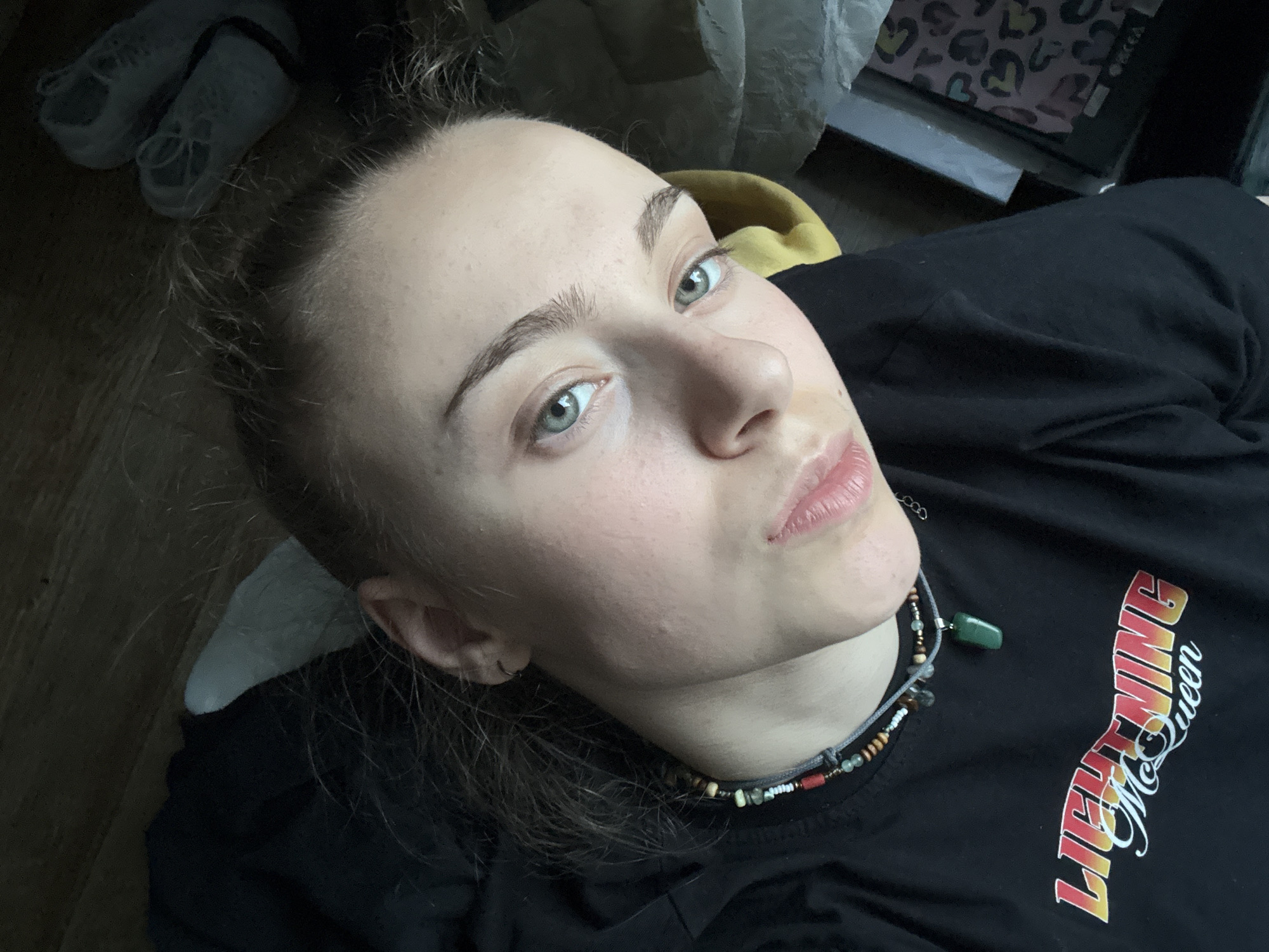 A young woman with light skin, light blue eyes, and light brown hair pulled back in a messy bun is positioned at an upward angle in the frame. She wears a black t-shirt with the phrase "Lightning McQueen" printed in white, red, and yellow letters on the right side. She has a beaded necklace with a small green stone pendant. A pair of white sneakers sits in the upper left corner of the frame, and a grey and black object with heart patterns is in the upper right. The background is out of focus. There is no clear indication that this photo was taken in Sofia, Bulgaria.
