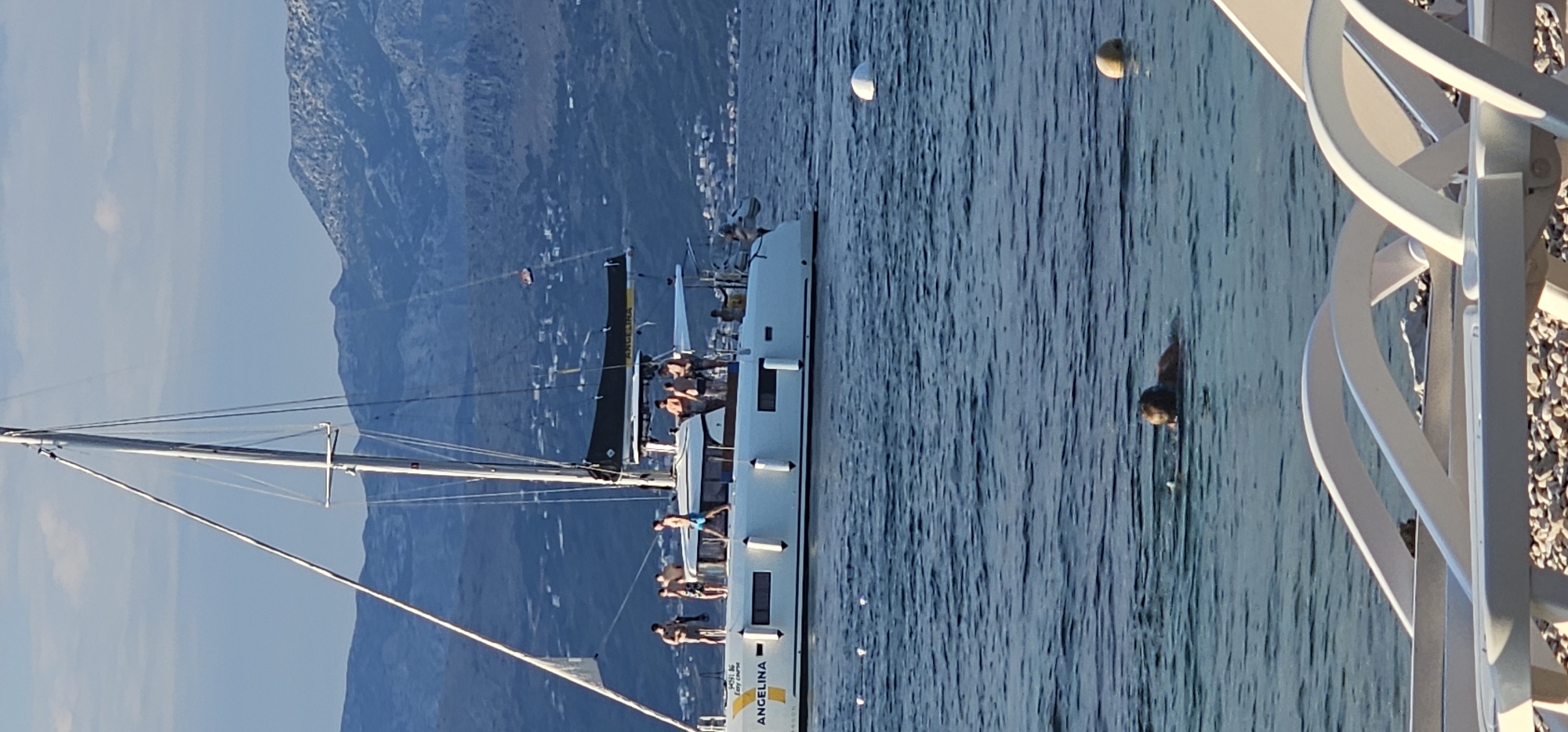 In the city of Supetar, Croatia, on a sunny day, a white catamaran named "ANGELINA" is anchored in the clear blue water. Several people are on the deck of the boat, some appearing to be sunbathing or socializing. In the foreground, a person is swimming in the sea, with lounge chairs visible on the shore in the very bottom of the frame. The background is dominated by a hazy, mountainous landscape, likely the island of Brač itself, with a scattering of buildings visible along the coastline. The sky is a light blue with a few wispy clouds.