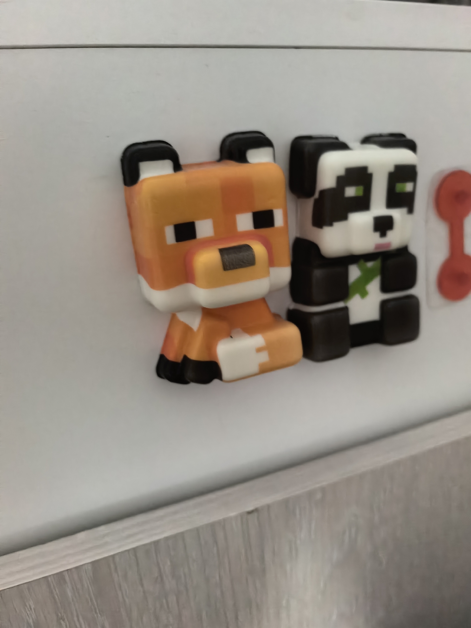 The image shows two Minecraft character magnets attached to a white surface. The magnet on the left is a fox, colored orange with black ears and blocky facial features. The magnet on the right is a panda, colored black and white with green eyes and a green cross on its chest. To the right of the panda magnet is a red magnet. The surface to which the magnets are attached appears to be a refrigerator or cabinet. The bottom of the image shows a light brown, textured surface, possibly a countertop or flooring. There is no indication of the image being in Sterlitamak, Russia.
