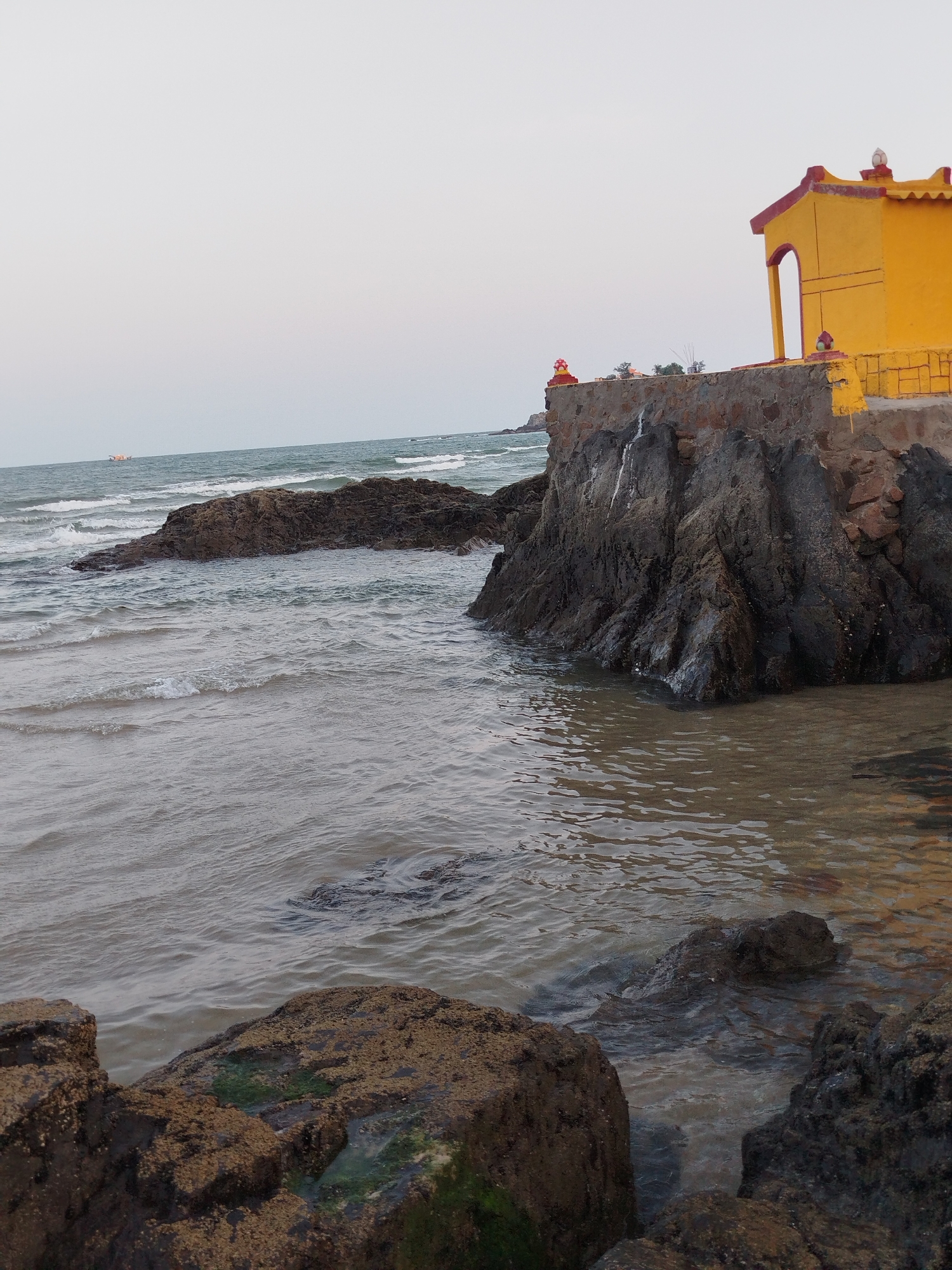 The image shows a rocky beach with a yellow building on a small cliff. The water is calm with small waves. There are rocks in the foreground and a larger rock formation further out. A boat is visible in the distance. The location is Ho Chi Minh City, Vietnam.