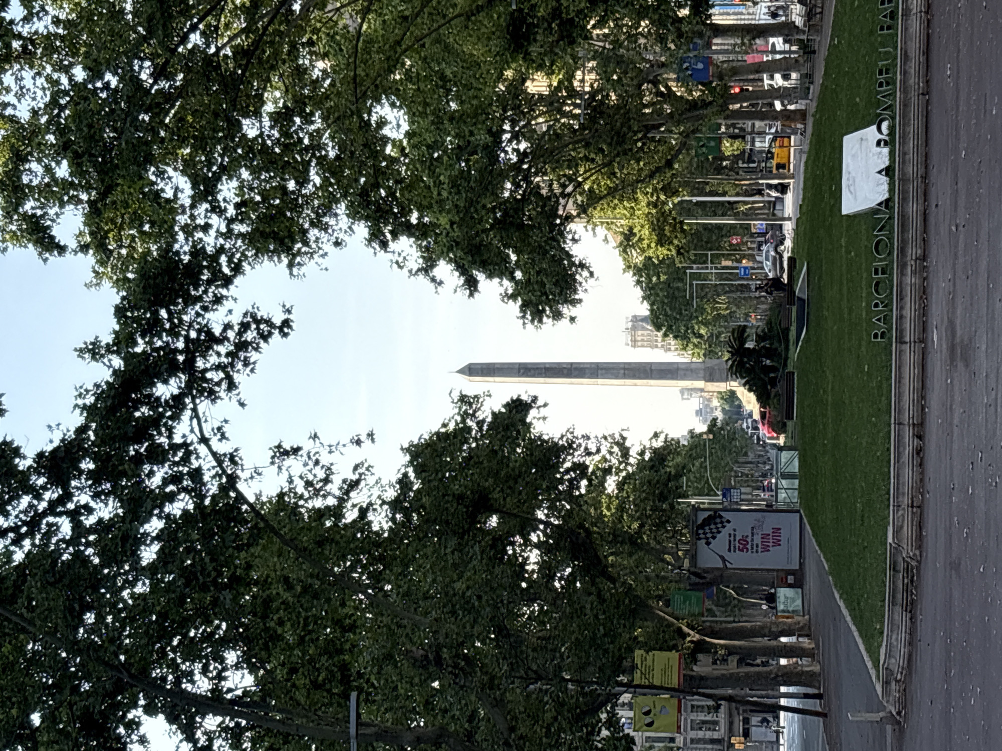 A street view in Barcelona, Spain, framed by trees on both sides, reveals a tall, slender monument in the distance. The monument, likely a commemorative pillar or obelisk, stands prominently against the skyline. Along the right side of the street is a grassy median with the words "BARCELONA A. POMPEU FABRA" displayed on a white sign. Street signs, advertising posters, and streetlights line the sides of the road. Vehicles, including cars and buses, can be seen in the distance, suggesting moderate traffic. The trees' canopy creates a partial enclosure, filtering the light and adding depth to the composition.