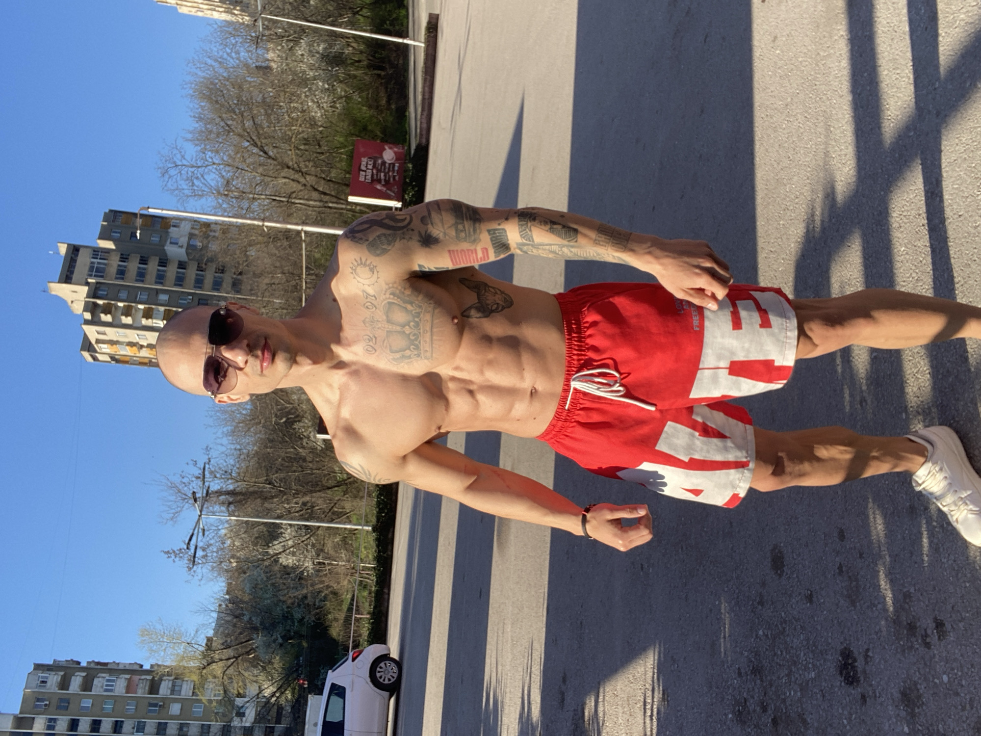 A shirtless man with tattoos is standing on an asphalt surface in Pleven, Bulgaria. He is wearing red and white shorts and white sneakers, and he is facing the camera with sunglasses on. In the background, there are buildings, trees, a white car, and a street sign. The scene is illuminated by bright sunlight, casting shadows on the ground.