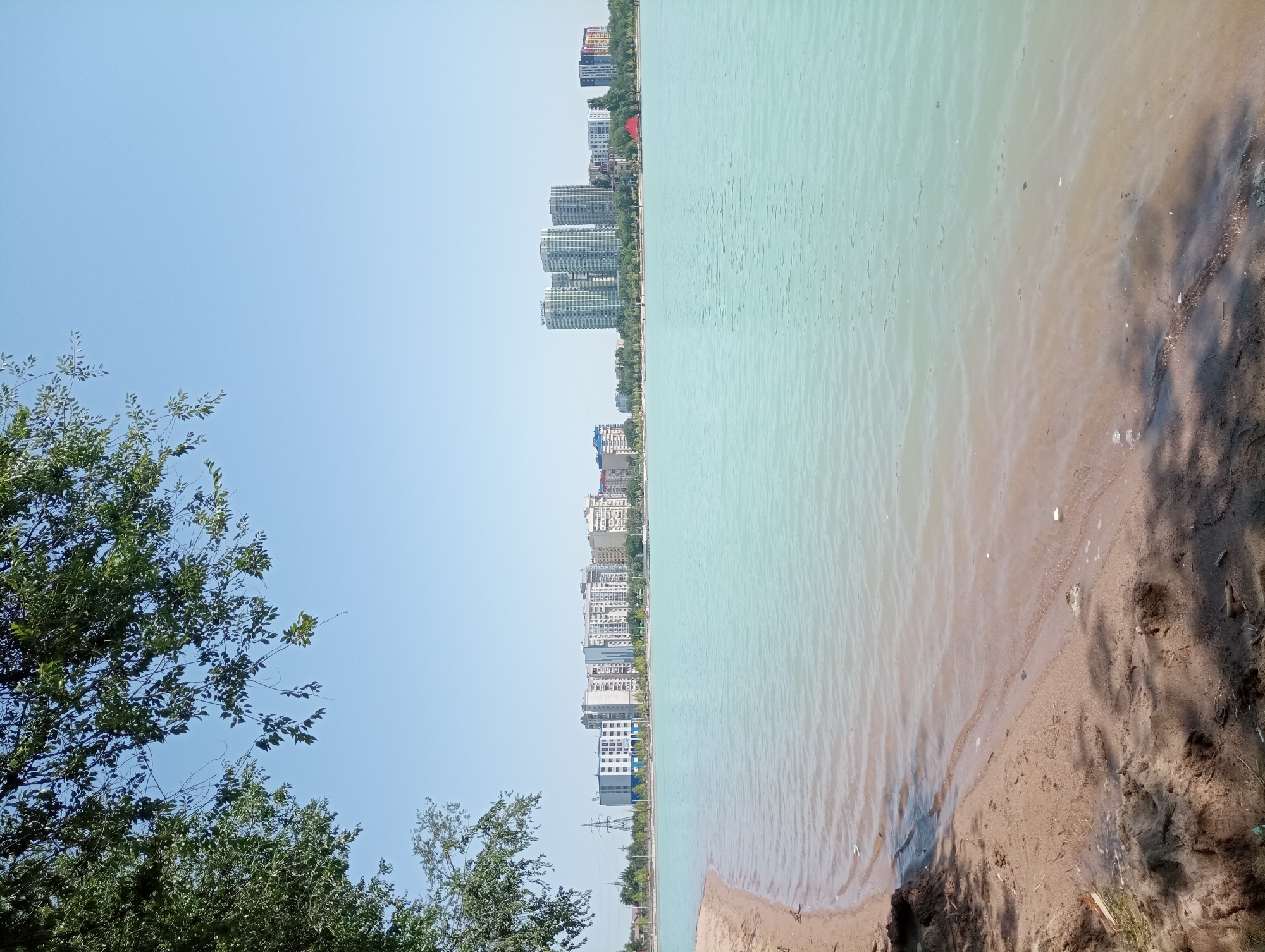 The image depicts a body of water, likely a lake, with a sandy shore in the foreground. The water is a light turquoise color and appears calm with gentle ripples. In the background, a city skyline is visible, characterized by a cluster of modern high-rise buildings. Trees and some lower-rise structures are interspersed between the buildings and the shoreline. The sky is clear and blue, indicating a sunny day. The scene suggests an urban waterfront environment in Almaty, Kazakhstan, possibly a recreational area or a developed part of the city with residential and commercial buildings.