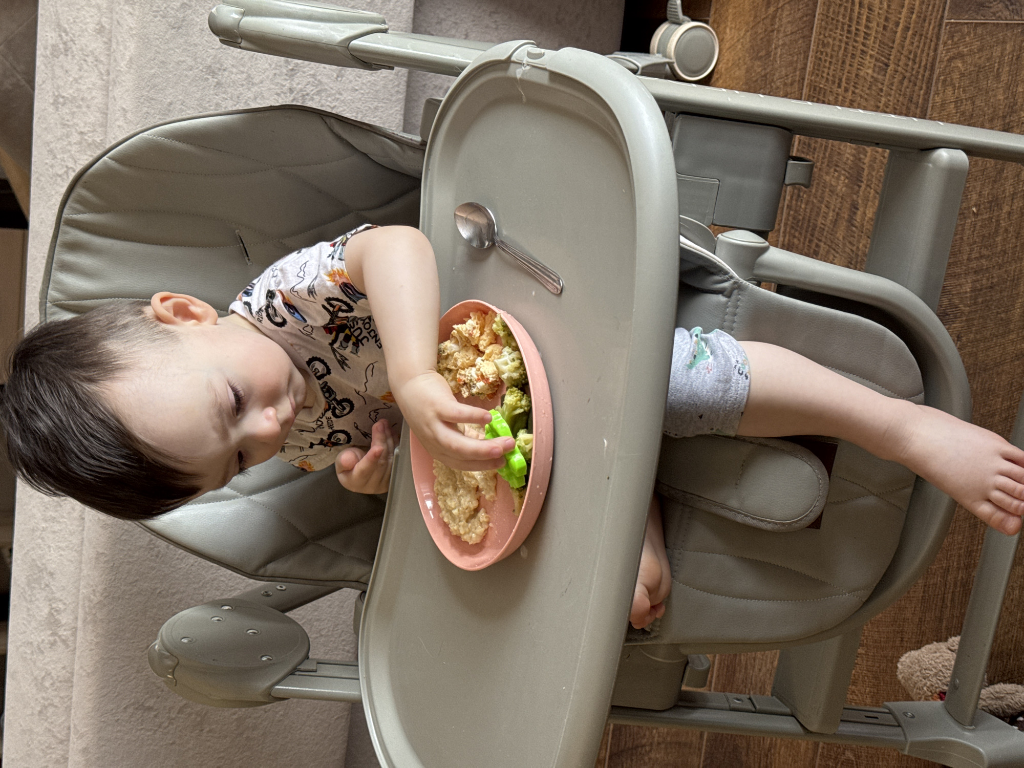 A young child, likely a toddler, with dark hair and light skin, is seated in a gray high chair during a mealtime. The child wears a white t-shirt patterned with vehicles and partially visible text including "ENJOY" and "RIDE," along with gray shorts. The child's head is tilted downwards, focused on a pink, possibly silicone, bowl of food. The left hand is reaching into the bowl, holding a piece of green vegetable, likely broccoli or cauliflower, mixed with a light-colored, soft food resembling oatmeal or cereal. A silver spoon rests on the gray high chair tray to the right of the bowl. The high chair features a padded seat and wheels on its frame. The setting appears to be indoors, likely a home, with a wooden floor visible and a light-colored, textured fabric, possibly a couch or curtain, in the background. A portion of a brown teddy bear is visible on the floor near the high chair's base. The activity shows the child in the process of eating, potentially self-feeding. The scene depicts a calm moment of a child's meal in Izhevsk, Russia.
