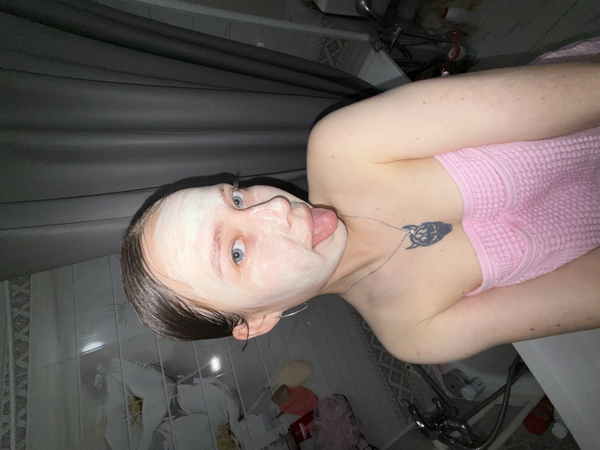 In this media file, a young person is depicted in what appears to be a bathroom. The subject has a white facial mask applied to their face and is sticking their tongue out, looking towards the camera with wide, blue eyes. They are wearing a pink towel wrapped around their chest and have a necklace with a pendant. A tattoo is visible on their upper chest.
The bathroom setting includes tiled walls with a decorative pattern and a dark shower curtain. Various bathroom accessories are scattered on the floor, including what looks like a loofah, a bottle, and possibly other hygiene products. The lighting suggests it could be nighttime or an indoor setting with artificial light. The overall mood seems playful and candid, with the subject engaging directly with the viewer.
There is no visible text in the image. The location context provided is Chaikovsky, Russia.