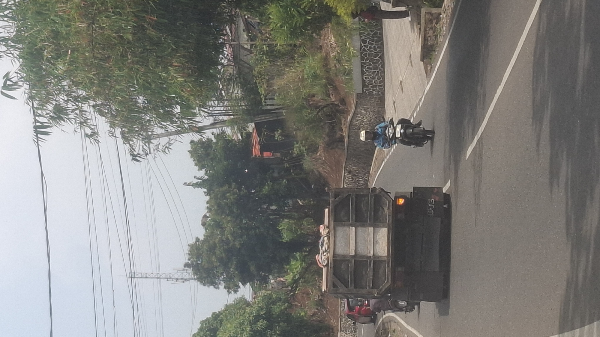 A low-angle, vertical, eye-level shot captures a street scene in Sirnagalih, Indonesia. The foreground is dominated by the rear of a large, dark-colored dump truck with "UTE" visible on its back. The truck is carrying a load of what appears to be bundled materials. To the right of the truck, a person wearing a blue jacket and a white helmet rides a dark-colored motorcycle forward. Further to the right and slightly behind the motorcycle, a person in a dark outfit stands on a sidewalk. The road itself is asphalt, marked with white lines. A stone wall borders the right side of the road. The background features lush green trees and foliage, with some houses visible among them. Power lines and a cell tower are visible in the hazy sky above. The overall atmosphere is that of a typical day on a semi-urban road.