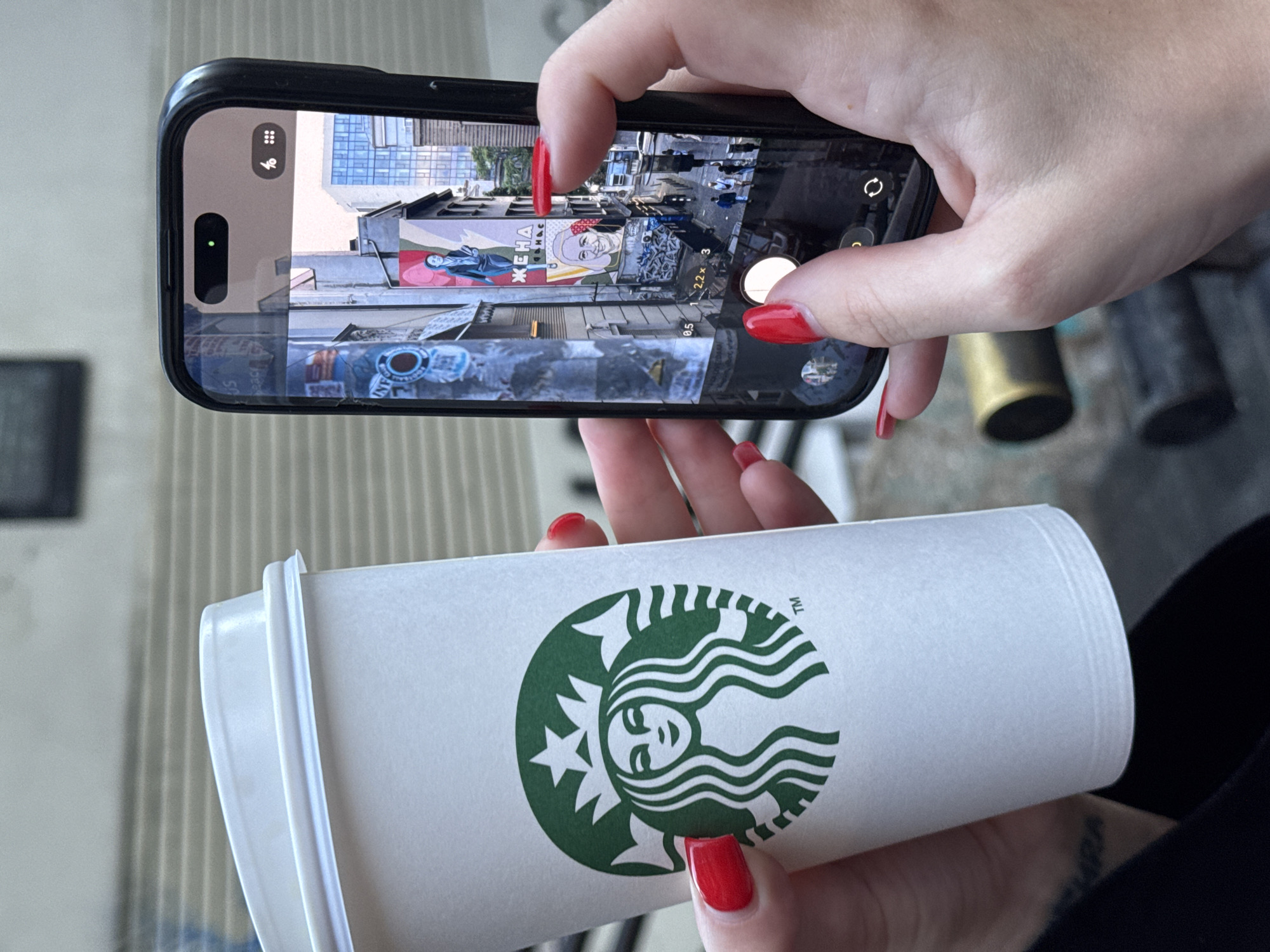 A person with red nail polish holds a Starbucks coffee cup in the lower foreground while simultaneously using a smartphone to photograph a wall mural in Belgrade, Serbia. The mural depicts a stylized image of a woman with the Cyrillic word "ЖЕНА" (meaning "woman") to the left of her portrait. The phone is held at eye level to frame the mural. The background shows city infrastructure including a building with glass panels.