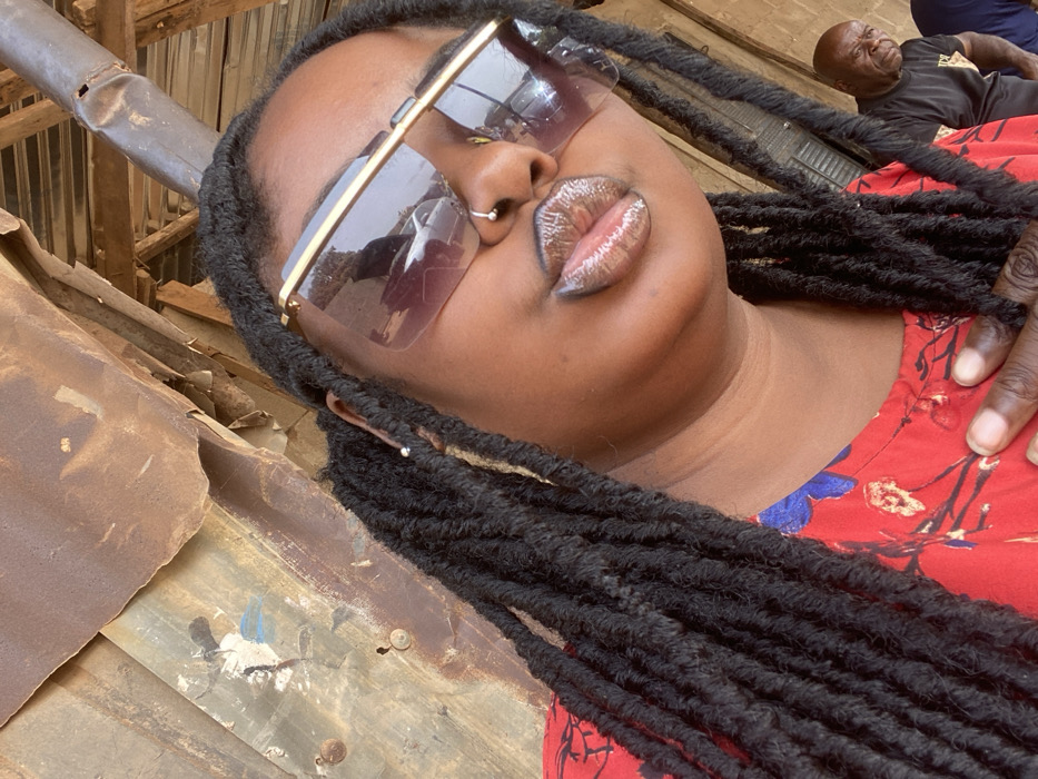 The image presents a close-up of a woman with an older man visible in the background, set in what is identified as Hayin Banki, Nigeria. The primary subject is a woman with dark skin and long black dreadlocks, positioned centrally and looking slightly upward and to her left. She wears large, rimless rectangular sunglasses with a gold-colored upper frame. A small silver ball stud pierces her left nostril, and a small silver stud is visible in her left ear. Her full lips are slightly pursed and appear glossy. She is dressed in a red garment featuring a blue and light-colored floral pattern. Her right hand, with a dark substance resembling nail polish or henna on the thumb, is placed against her chest area. In the lower right background, an older man with dark skin and a bald head is partially visible, wearing a dark t-shirt, looking toward the right edge of the frame with a neutral expression. The background consists primarily of weathered, rusted corrugated metal sheets, with visible fasteners and patches of faded blue and white paint. Wooden structural beams are evident above and behind the subjects, suggesting a rustic or makeshift overhead cover or shelter. Reflections in the woman's sunglasses show blurred images of a street scene, including what appears to be a white vehicle.