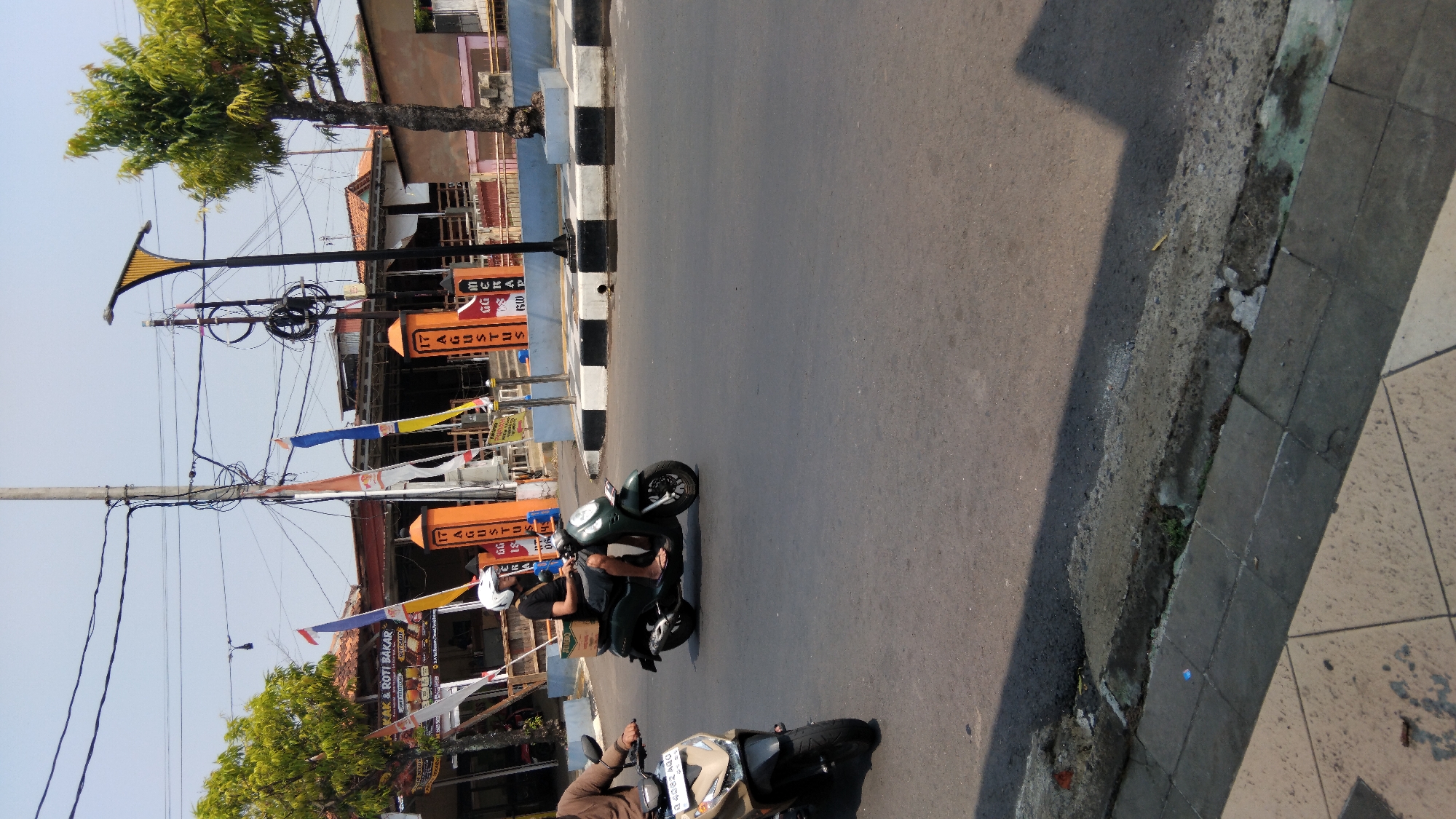 In Batang, Indonesia, a man on a dark green scooter is riding on a paved road. He is wearing a white helmet and a black t-shirt. The scooter has a brown box attached to the back. Another scooter, beige and black, is visible to the left, with its rider partially out of frame. Behind the scooters, a building with orange pillars and flags is visible. These pillars are decorated with vertical text that appears to be in Indonesian. Trees line the street, and power lines crisscross overhead. The sky is clear and bright, indicating a sunny day. The road surface is asphalt, with a sidewalk composed of square tiles on the left side of the frame.