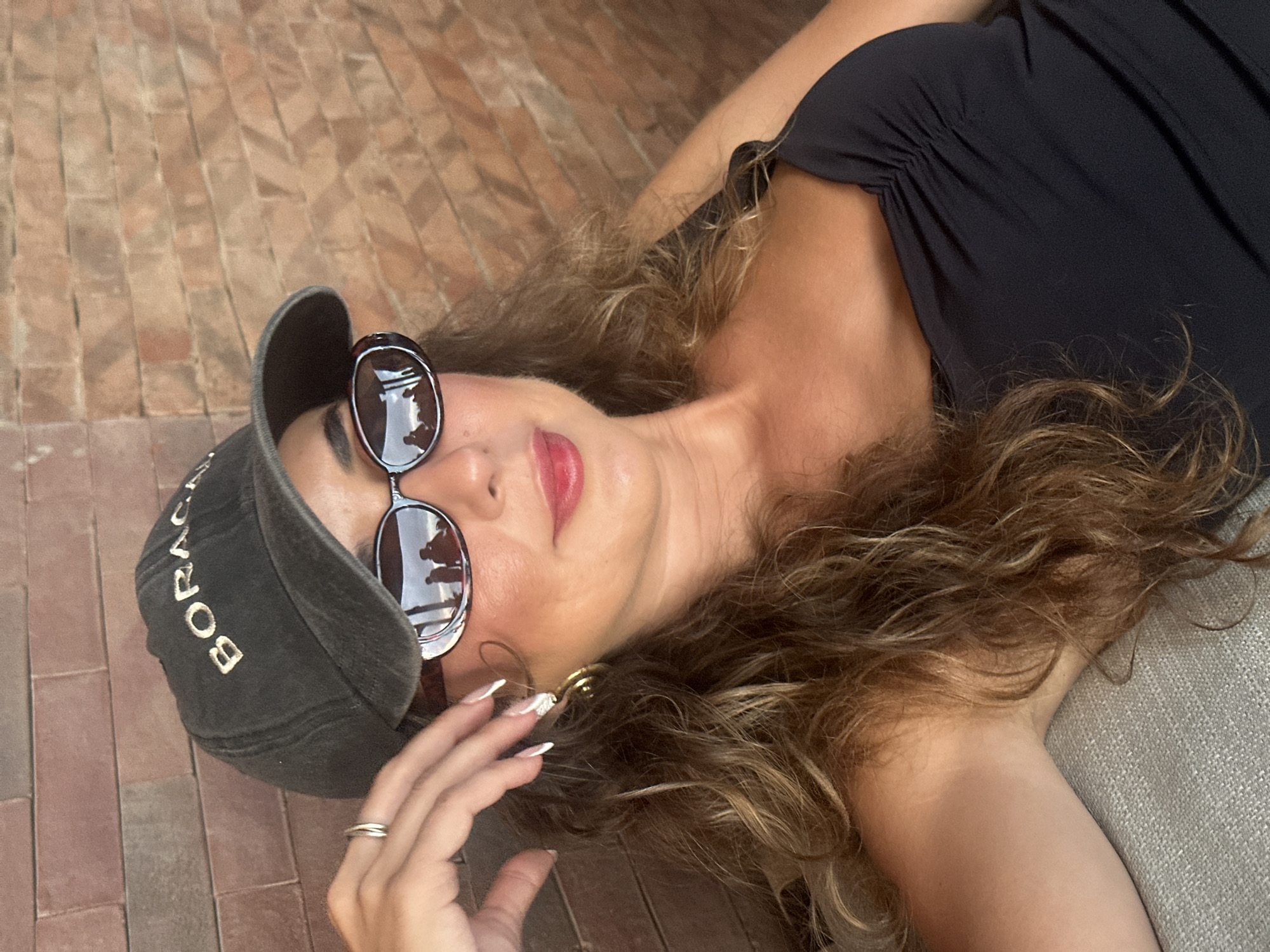 A woman is lying down, wearing a black cap with the text "BORACA", oval sunglasses, a black top, and a ring on her left ring finger. She is also wearing gold earrings and has long, curly brown hair. The background includes brick paving. The reflection in the sunglasses appears to show people and the location. The scene is located in North Kuta, Indonesia.