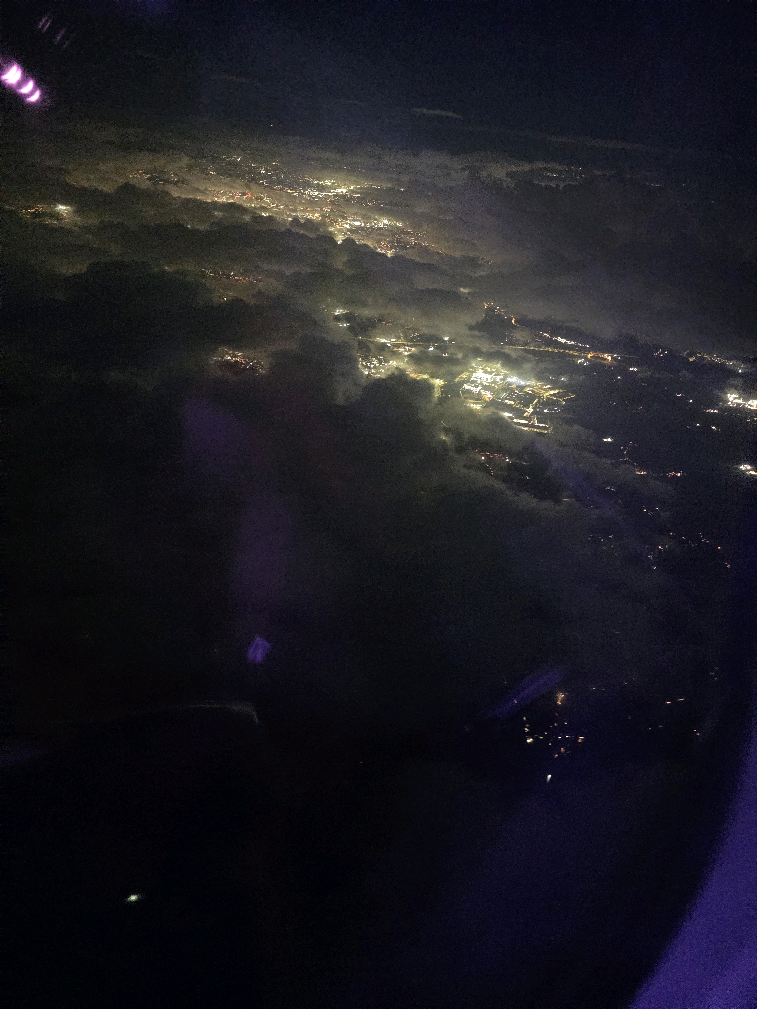An aerial photograph taken at night shows a sprawling urban landscape partially obscured by clouds. Numerous artificial lights, indicative of a city, illuminate the scene below. Some lights appear clustered together, suggesting densely populated areas or industrial zones, while others are more dispersed. The clouds create a textured overlay, with some areas completely concealing the ground and others allowing glimpses of the lighted city. There are also lens flares visible, manifesting as purple and white streaks of light across the upper left and lower right portions of the image. The overall impression is of a vast, brightly lit city seen from a high vantage point above the clouds. The specific location within North Warwickshire, United Kingdom, cannot be determined from the visual information provided in the image.