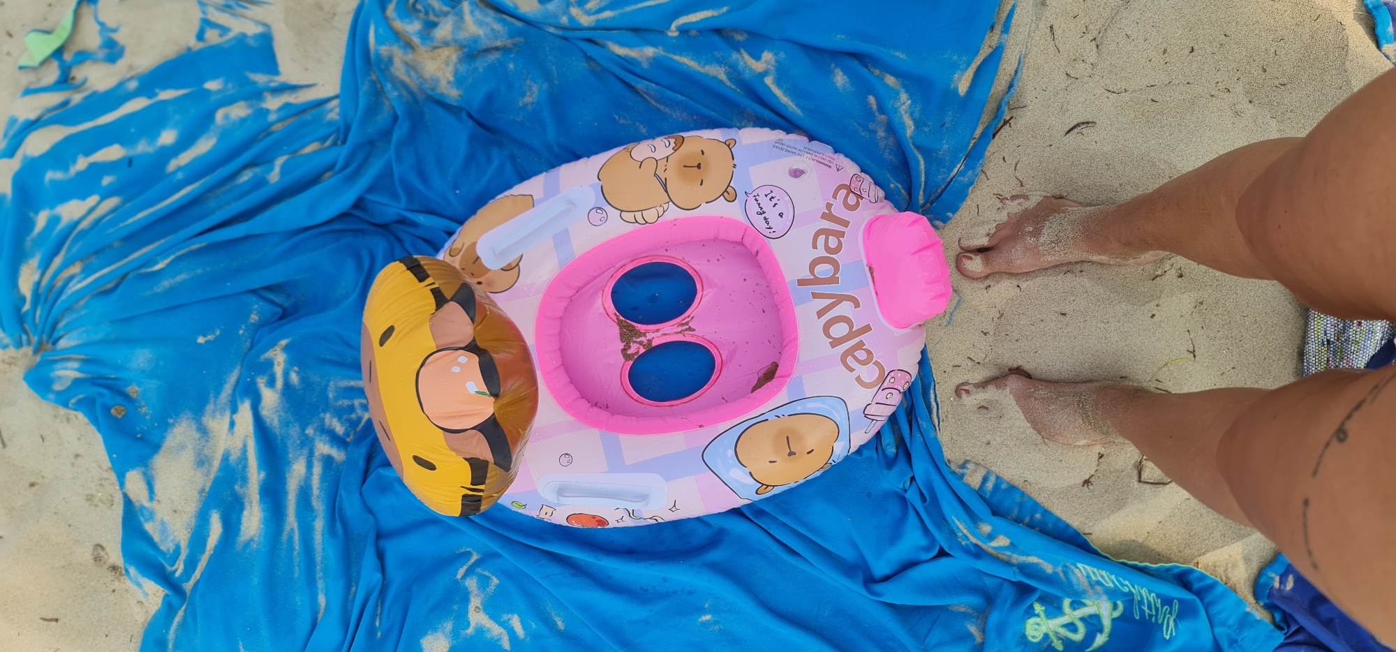 The image shows a beach scene, likely in Đà Nẵng, Vietnam given the sandy shore. A person is positioned near the right edge of the frame, their legs and feet visible with some sand covering them. A bright blue cloth covers a large portion of the image, possibly a beach towel or sarong. On the cloth sits an inflatable baby floatie. The floatie is pink, tan, and yellow, and features cartoon bear designs and a pink seat with two holes. Sand is visible on the cloth and the floatie, as well as in the background which appears to be the beach. The overall impression is a relaxed beach setting.