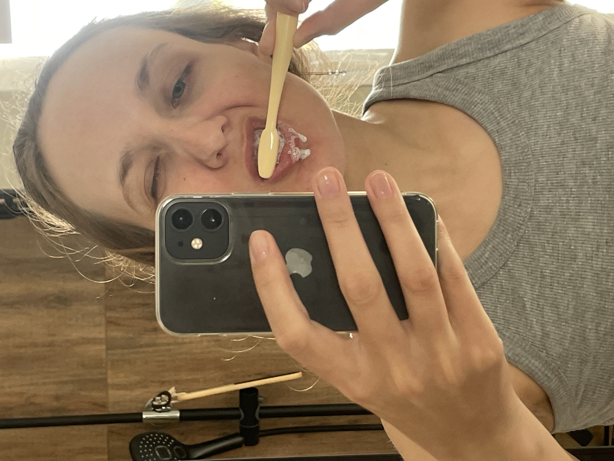 A person is taking a selfie while brushing their teeth with a yellow toothbrush. The person is wearing a gray shirt. They are holding a black iPhone in front of their face. The background shows a wooden wall and a black object on a wooden surface. There is no evidence to suggest the location is Подшивалово, Russia.
