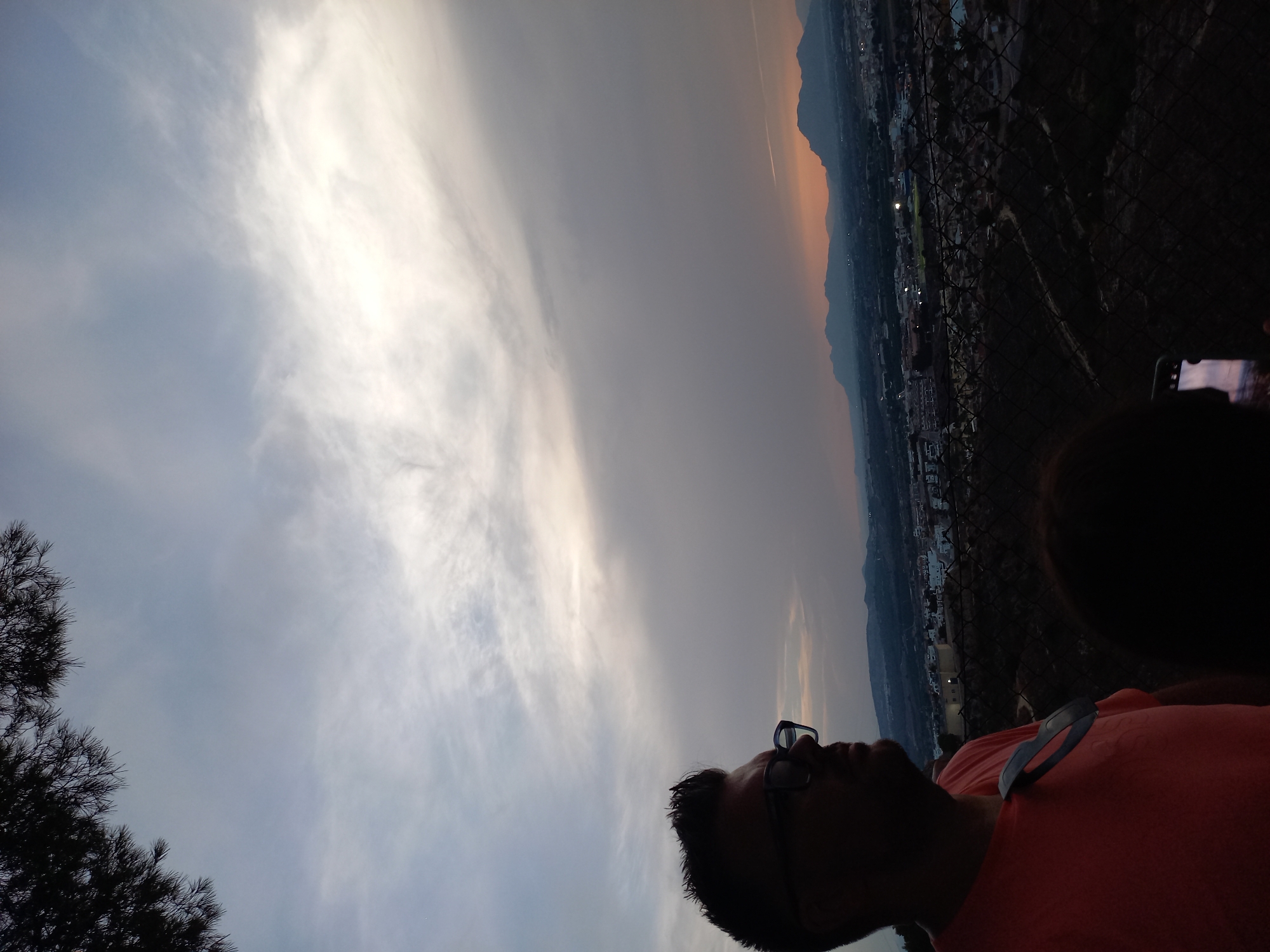 A silhouette of a man wearing glasses and an orange t-shirt is visible in the foreground, looking out at a panoramic view of Urbanización Ciudad Quesada, Spain. The sky is overcast with hints of sunset colors peeking through. The cityscape stretches out below, dotted with lights, and is framed by rolling hills in the distance. A dark fence is in the middle ground.