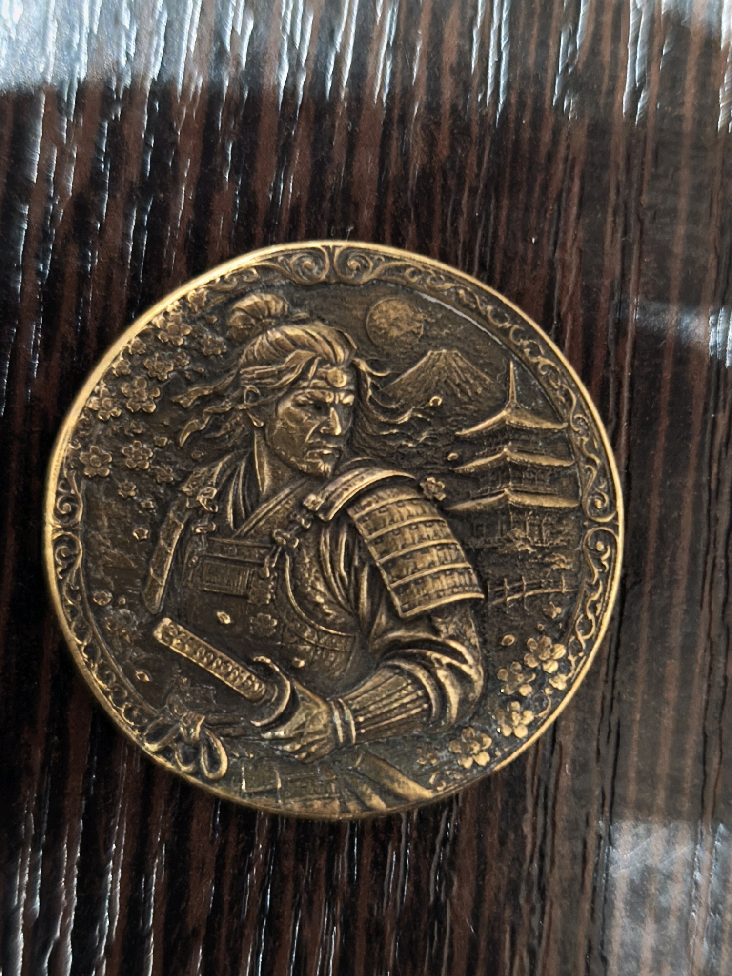 The image displays a close-up, high-angle shot of a round, antique-looking medallion. The medallion is made of a dark, metallic material, possibly brass or bronze, with intricate engravings. The scene depicted on the medallion features a samurai warrior in full armor, facing left. The warrior has a stern expression, with long, flowing hair and a headband. He is holding a sword in his left hand. In the background, there is a traditional Japanese pagoda and Mount Fuji under a crescent moon. The medallion is placed on a dark, textured surface that appears to be wood with a prominent grain pattern. The lighting is dim, casting shadows that highlight the relief of the medallion's design. The context of Karachayevsk, Russia, is not visually evident in this image.