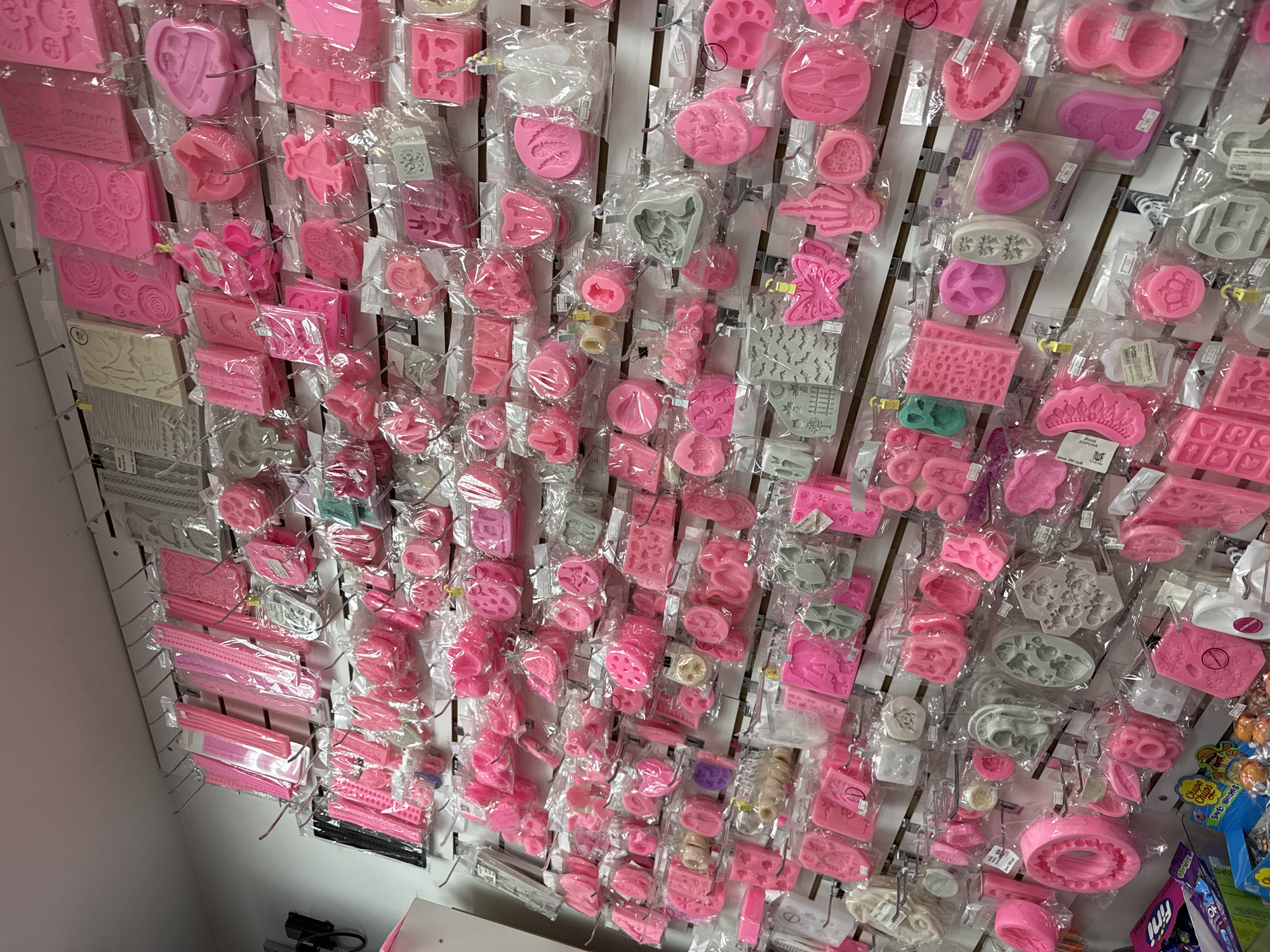 The image depicts a brightly lit retail store interior, likely a bakery supply or craft store, filled with an extensive display of silicone molds. The molds are predominantly pink and vary in shape and size, featuring intricate designs such as flowers, animals, geometric patterns, and holiday-themed items. They are neatly organized and hung on a pegboard wall, with many individually packaged in clear plastic. The store appears well-stocked, with shelves extending from floor to ceiling. The overall impression is one of a specialized retail environment focused on baking and crafting supplies. No specific location details other than the city of Lyubertsy, Russia, are visible or inferable from the image. There are no people or actions depicted in the image.