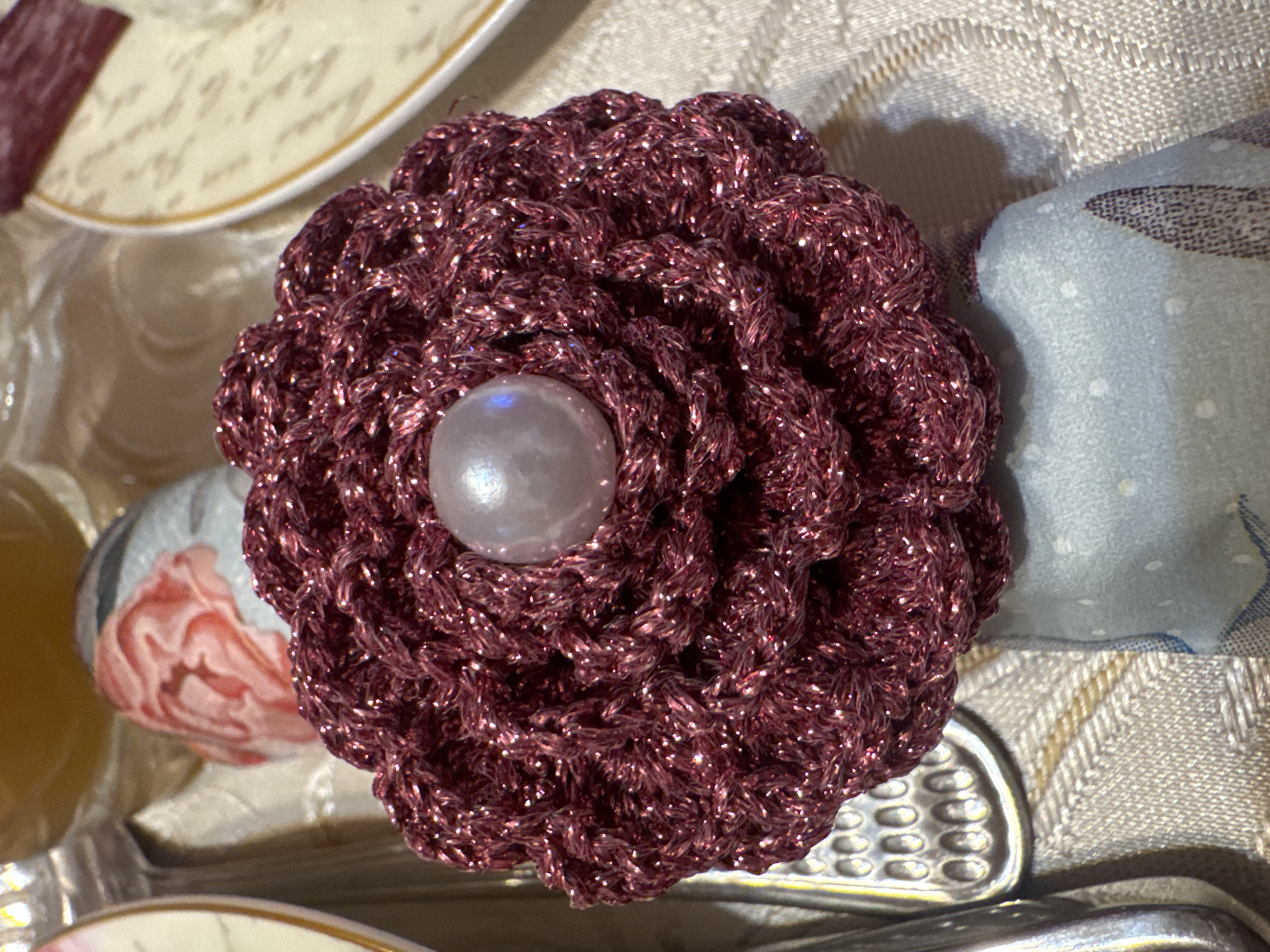 A close-up photograph shows a crocheted flower, burgundy in color with sparkling threads, centered on a light-colored tablecloth with a subtle woven pattern. The flower has a large, pearly-white bead at its center. Fragments of dishware appear in the periphery: a plate with patterned text and gold rim, and a floral-patterned decorative item. Cutlery, possibly a fork or serving spoon, is visible beneath the flower. A soft, blue and white patterned object rests against the right edge of the flower. The photo gives no indication of the location in Plovdiv, Bulgaria.
