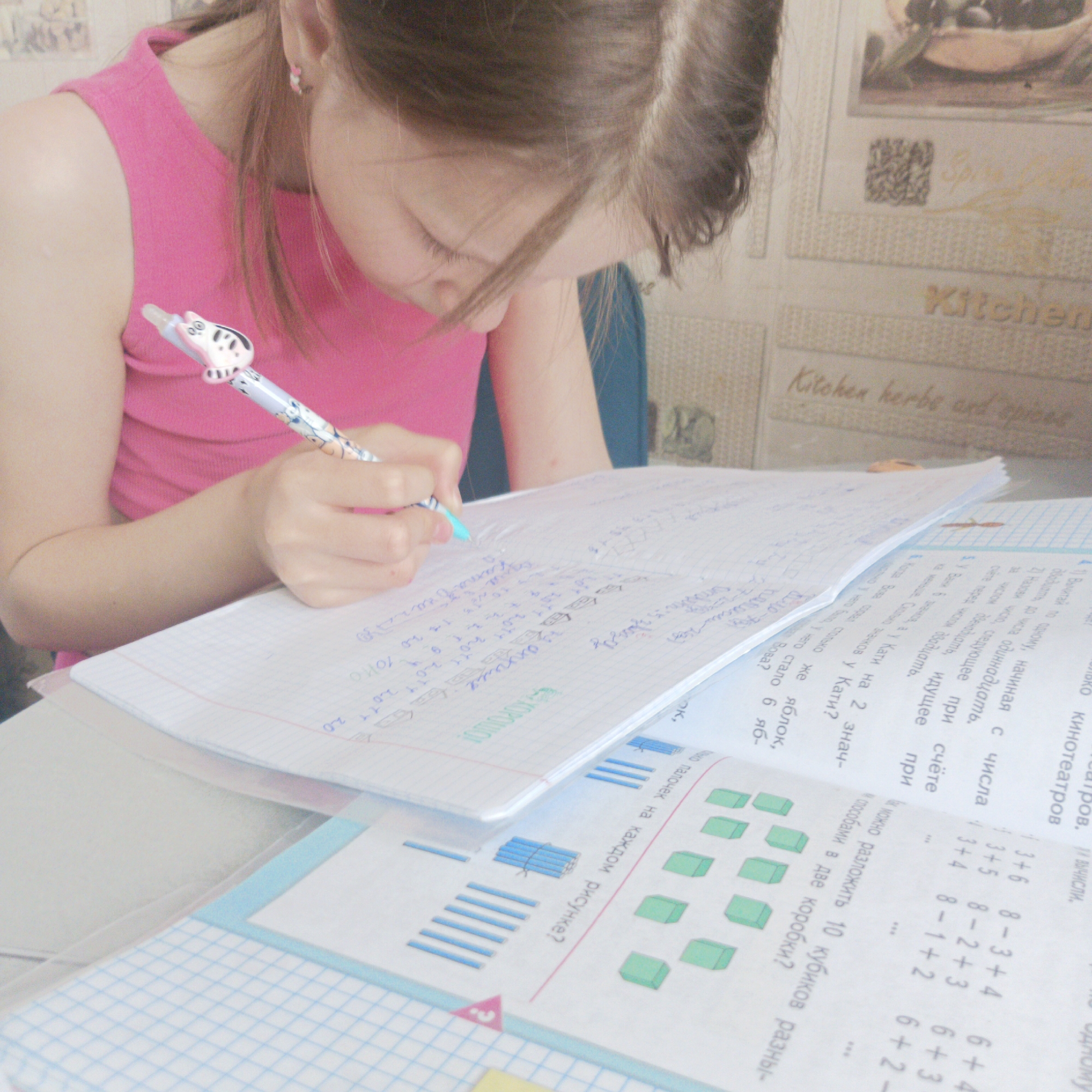 A young girl with brown hair, wearing a bright pink sleeveless top and a small earring, is intently focused on writing in a notebook. She holds a light-colored pen, adorned with a white and black cat-like topper, as she bends over her schoolwork. The scene captures her in a moment of concentration, likely doing homework or engaging in a study session indoors during the daytime.
On the table in front of her, an open notebook filled with handwritten mathematical equations and notes in blue and green ink rests atop a workbook. The workbook displays various math problems and illustrations, including blue sticks and green cubes, likely used for counting exercises. Visible text in Russian on the workbook includes instructions such as "Вычисли." (Calculate.) and word problems like "У Вани 6 значков, а у Кати на 2 знач- ка ... Сколько значков у Кати?" (Vanja has 6 badges, and Katya has 2 badges... How many badges does Katya have?), as well as numerous arithmetic operations like "3+6", "8-3+4", and "6+4".
The background features wallpaper with a kitchen theme, bearing the words "Kitchen" and "Kitchen herbs and spices" in decorative script. An illustration of olives in a bowl is also visible on the wallpaper. These details suggest the setting is a kitchen in a home, possibly in Chaikovsky, Russia, as indicated by the location context. The overall impression is one of a diligent child engaged in learning.