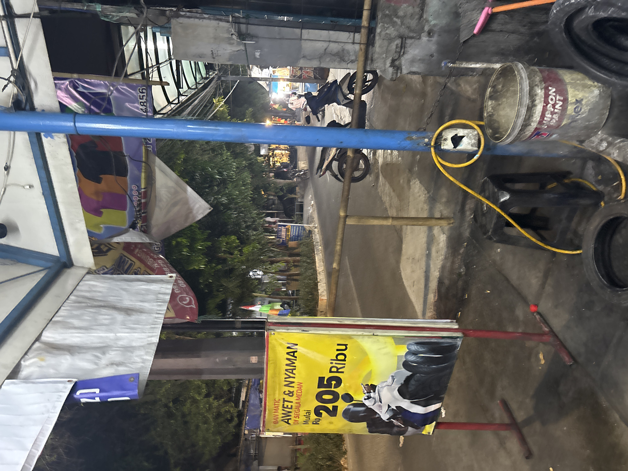 In North Jakarta, Indonesia, an outdoor scene is captured at night, likely on a street or service area. A bright yellow advertisement sign stands prominently on the left, featuring a picture of a motorcycle and a price of "Rp 205 Ribu" (205,000 Rupiah), indicating a sale or service. Above the price, text reads "BAN MATIC AWET & NYAMAN DI SEGALA MEDAN" (Matic Tires, Durable & Comfortable in All Terrains).
A blue pole, part of an overhead structure or utility pole, stands near the center of the image, supporting various wires. A yellow electrical cord is wrapped around the pole, attached to a piece of white material with a black symbol. To the right of the pole, a black stool sits on the ground, and further to the right, a weathered bucket labeled "NIPPON PAINT" is visible. Partially obscured by the bucket and pole are two tires on the ground, one of which appears to be a spare.
In the background, a motorcycle is parked on the asphalt road. Beyond the road, there are trees and buildings, with some lights illuminating the area, suggesting shops or residences. A small Indonesian flag is visible on a pole in the distance. The overall environment suggests a commercial or roadside service area operating into the evening.
