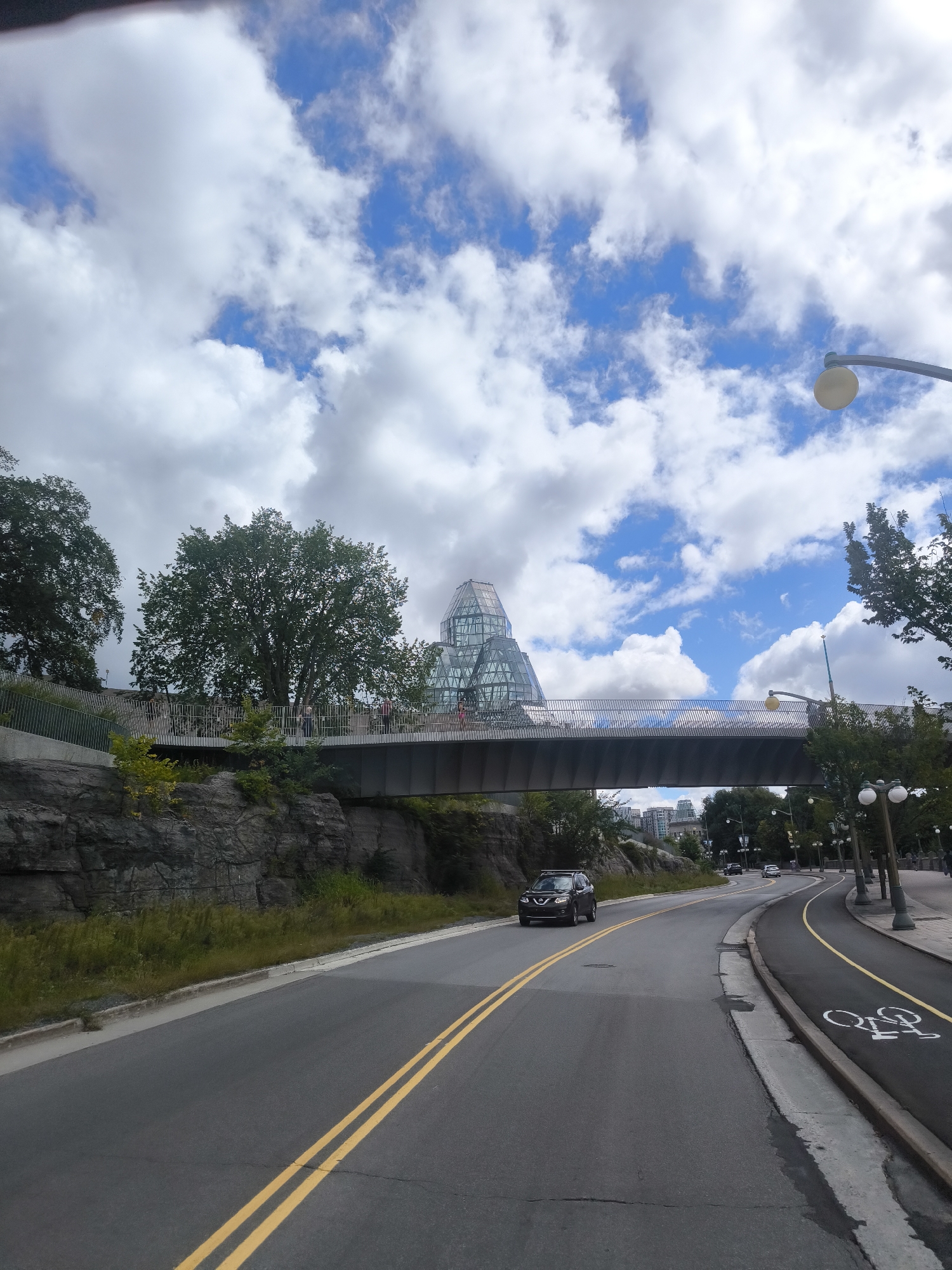 This is a street-level view looking towards a pedestrian bridge spanning over a road in Ottawa, Canada. The bridge is modern in design with a silver-colored railing and a dark grey deck. Beyond the bridge, a distinctive building with a glass-paneled, multi-faceted dome shape is visible, which is the National Gallery of Canada. On the left side of the road, there is a rocky embankment with some greenery. On the right side of the road, there is a bike lane marked with a bicycle symbol. A dark-colored SUV is driving on the road in the foreground, and a few cars are visible further down the road. The sky is partly cloudy with patches of blue sky. Trees line the sides of the road and the embankment. There appear to be a few people walking on the pedestrian bridge.