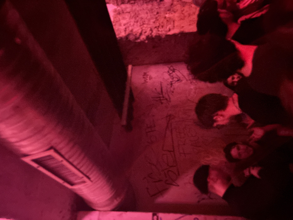 A dimly lit space with red hues reveals a group of people standing. Some have their backs to the viewer while others are facing forward. Their faces are partially visible in the subdued lighting. The backdrop features graffiti-covered walls. A large, cylindrical metal structure with a rectangular opening runs along the left side of the image, casting shadows and adding to the overall atmosphere. The composition is tilted, enhancing the sense of depth.