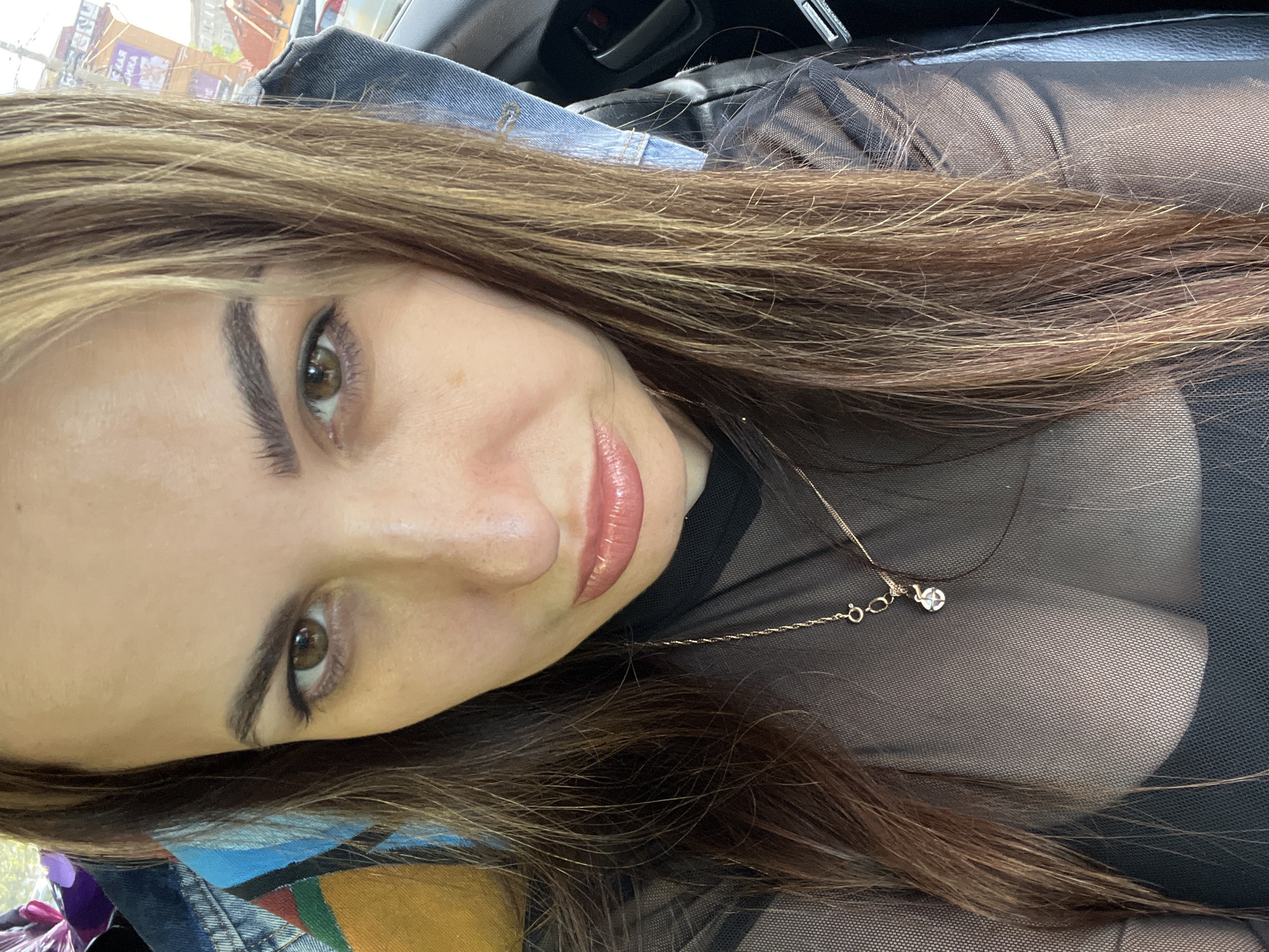 A selfie-style head and shoulders shot of a young woman with long, light brown hair. Her hair is parted off-center, and she wears a thin, delicate gold chain necklace with a single diamond or crystal pendant. The woman has fair skin, light brown eyes, and groomed eyebrows. She is wearing a black mesh top, beneath which can be seen the contours of her chest. The background suggests she is inside a car, with part of a denim jacket or shirt visible, along with parts of the car's interior. The overall setting appears to be urban, although specific details of the city are indiscernible.