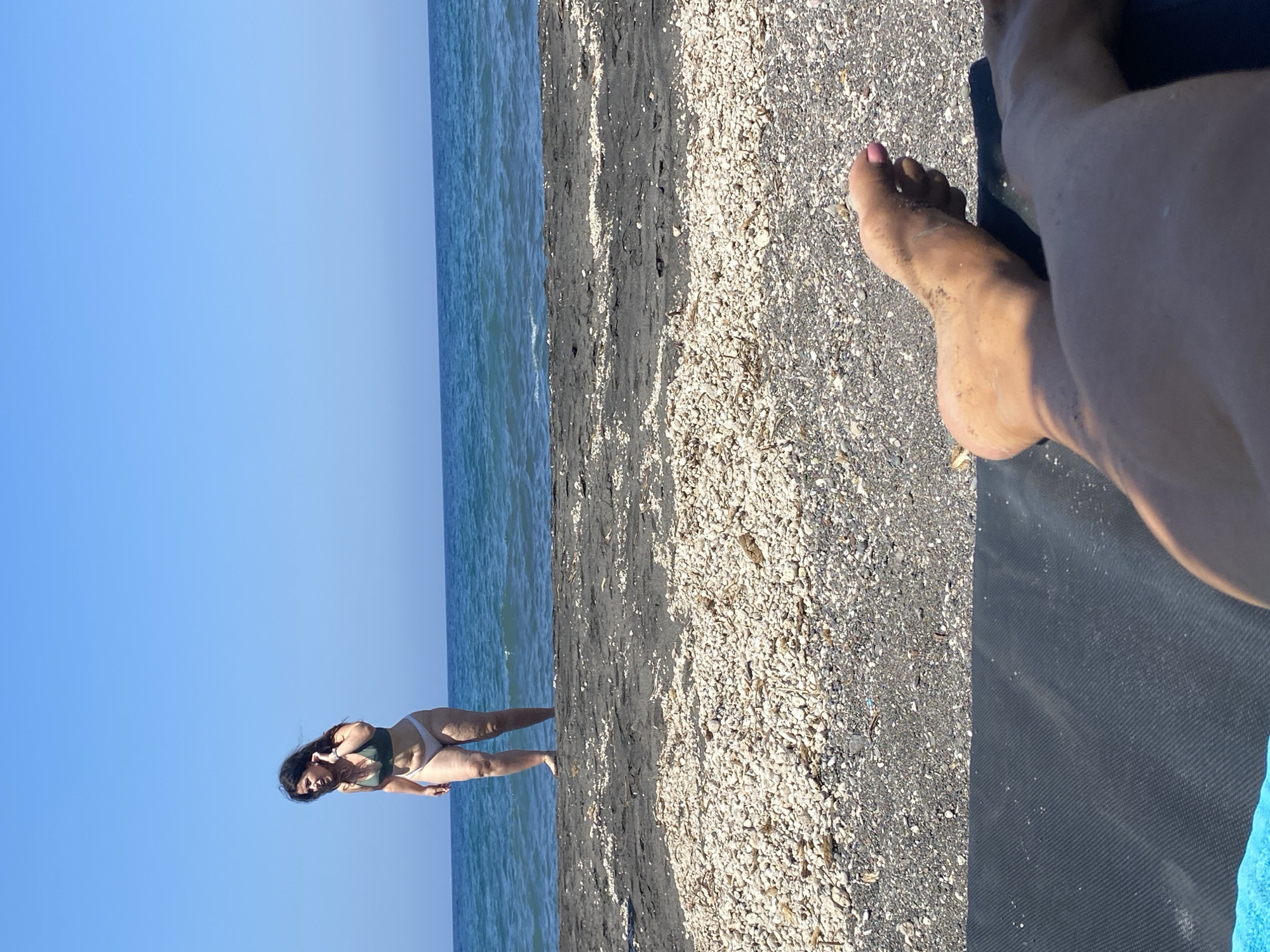 A woman in a dark green bikini is standing at the edge of the sea on a black sand beach in the city of Thira Municipal Unit, Greece. She is looking to her left and touching her hair. The ocean is calm and blue. The beach is covered in black sand and small white pebbles. In the foreground, a person's bare feet are visible, resting on a dark mat. The sky is clear and blue.