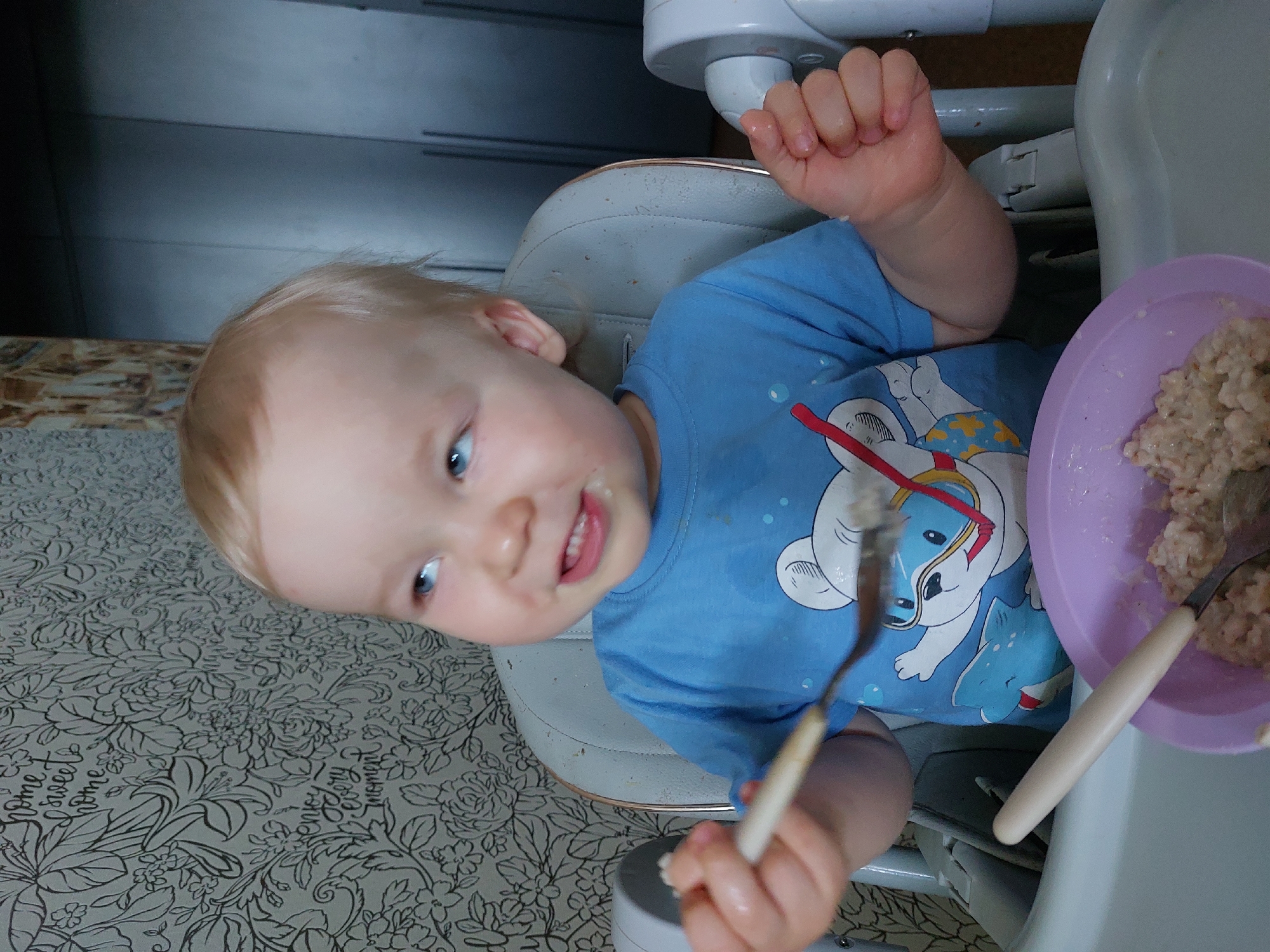 The image captures a young child, likely a toddler, seated in a high chair during a meal. The child has fair skin, light blonde hair, and bright blue eyes, looking towards the viewer with a soft, content smile. Some food residue is visible around their mouth, suggesting they are actively eating. They are wearing a blue t-shirt adorned with a cartoon polar bear design.
The child is in a light-colored high chair, with a purple plastic bowl of what appears to be oatmeal or porridge on the tray to their right. One spoon with a light handle is held loosely in the child's right hand, while another identical spoon is resting in the bowl.
The background behind the child is a dark, solid surface, possibly a wall or cabinet. Below and to the left of the high chair, a patterned surface is visible, featuring intricate black floral designs and text on a light gray background. Clearly visible phrases on this pattern include "home sweet home" and "every moment." The setting appears to be an indoor, domestic environment.