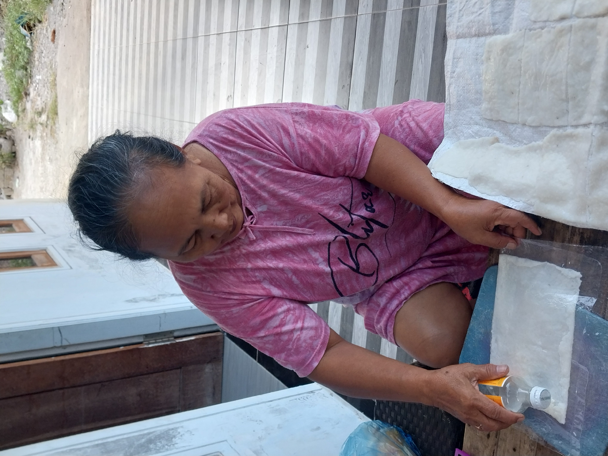 In Nglegok, Indonesia, an elderly woman wearing a pink tie-dye t-shirt is kneeling on a tiled porch, focusing on a process involving white, paper-like sheets and a small bottle of liquid. Her hands are engaged with these materials. One hand holds the bottle, seemingly about to pour its contents onto a sheet placed on a blue mat over a wooden surface. Another hand rests on the sheet. To her right, more white sheets are arranged on a textured white material. The background reveals a white building with wooden-framed windows, a wooden door, and some sparse vegetation and ground outside. The overall scene suggests a domestic, hands-on activity, possibly related to food preparation or crafting.